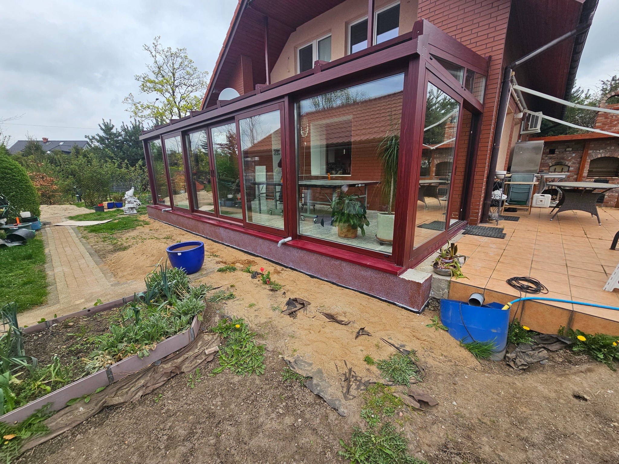 Zadaszony taras z dużymi oknami w brązowych ramach, widok z zewnątrz na ogród w trakcie prac ziemnych, z widocznym fragmentem domu z cegły.