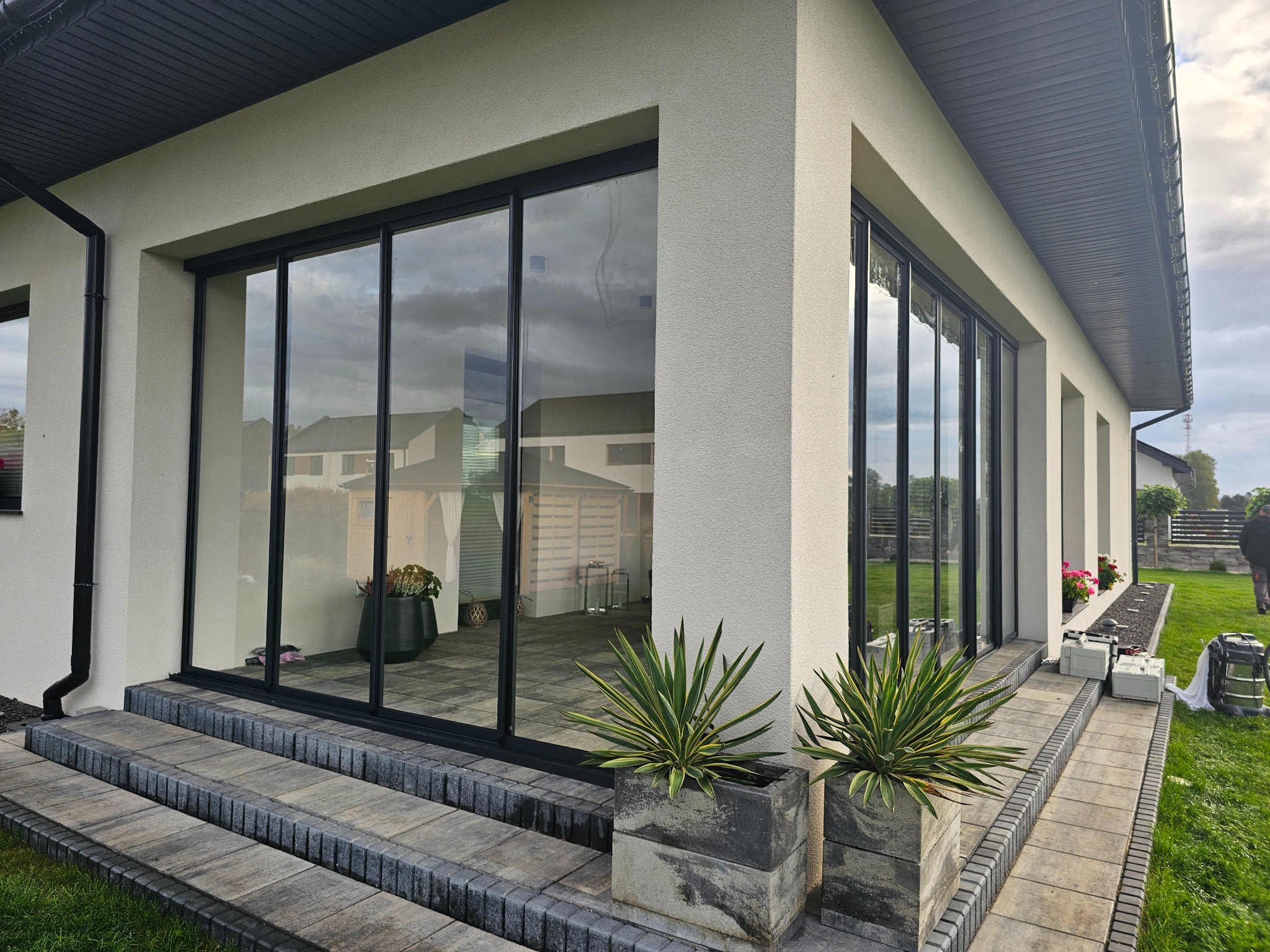 Nowoczesny dom z dużymi, przesuwnymi oknami z czarnymi ramami, widoczne dwustopniowe schody z szarej kostki brukowej i rośliny w betonowych donicach, w tle odbicie ogrodu w szybach.