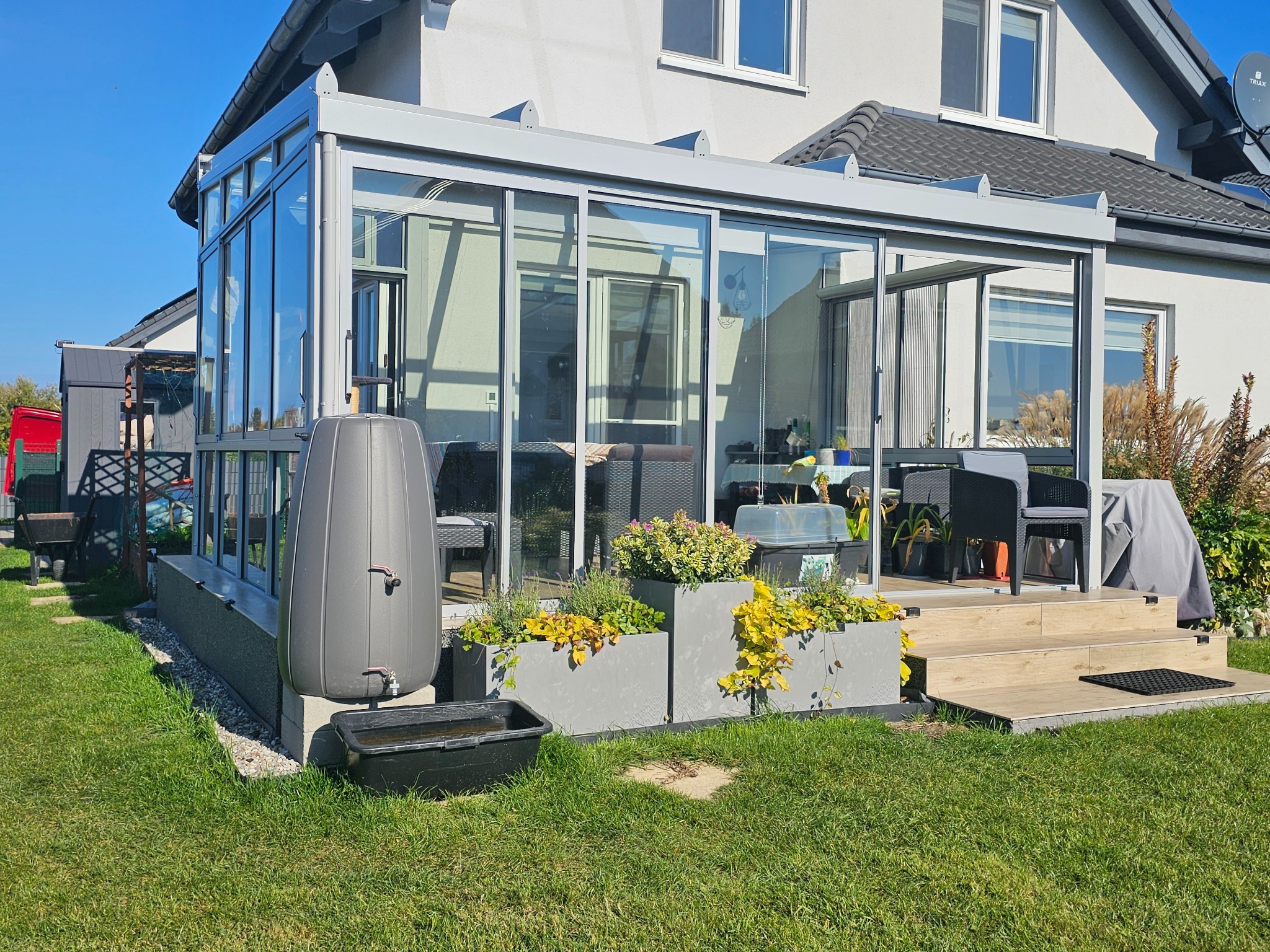 Szklana zabudowa tarasu z przesuwnymi drzwiami, widok z zewnątrz, z elementami dekoracyjnymi w postaci roślin w donicach i zbiornika na deszczówkę.