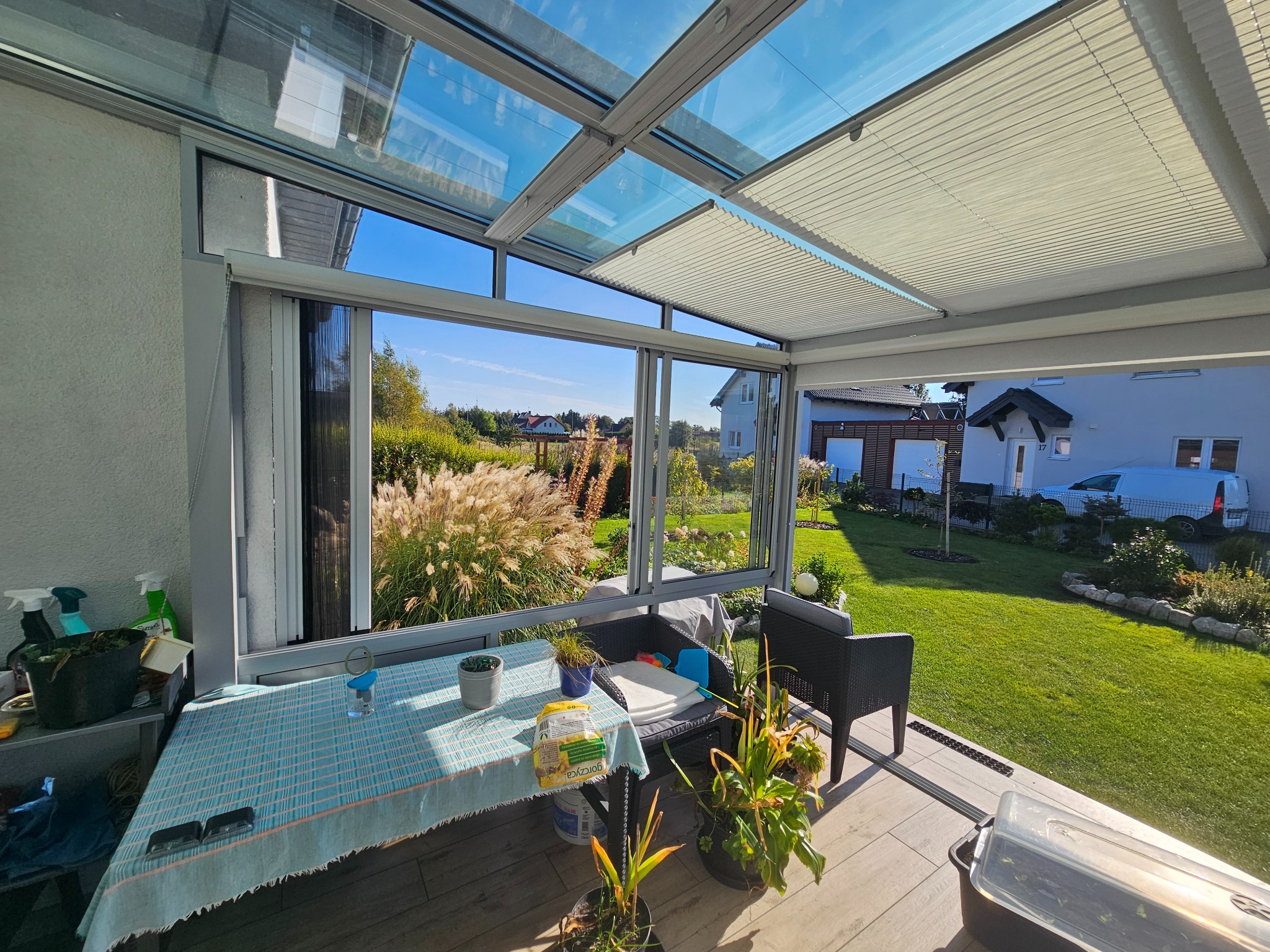Szklana zabudowa tarasu z aluminiową konstrukcją i roletami plisowanymi, widok na ogród z ozdobnymi trawami i domami w tle.