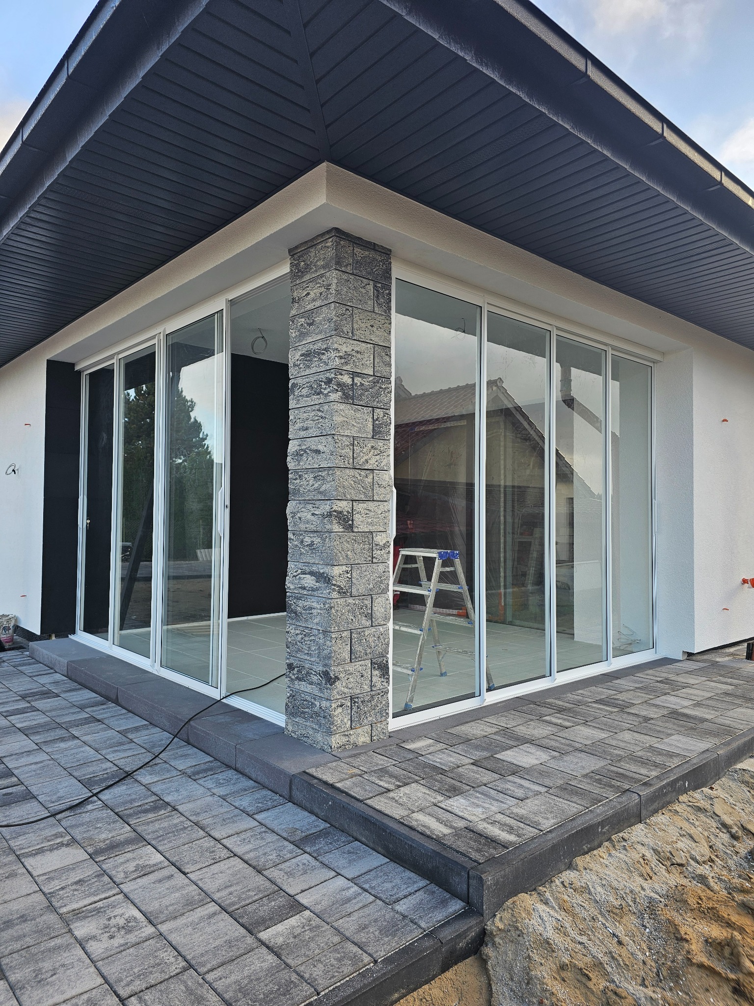 Nowoczesny dom z dużymi, przesuwnymi drzwiami szklanymi, wykończony kamienną kolumną i kostką brukową. W tle widoczna drabina.