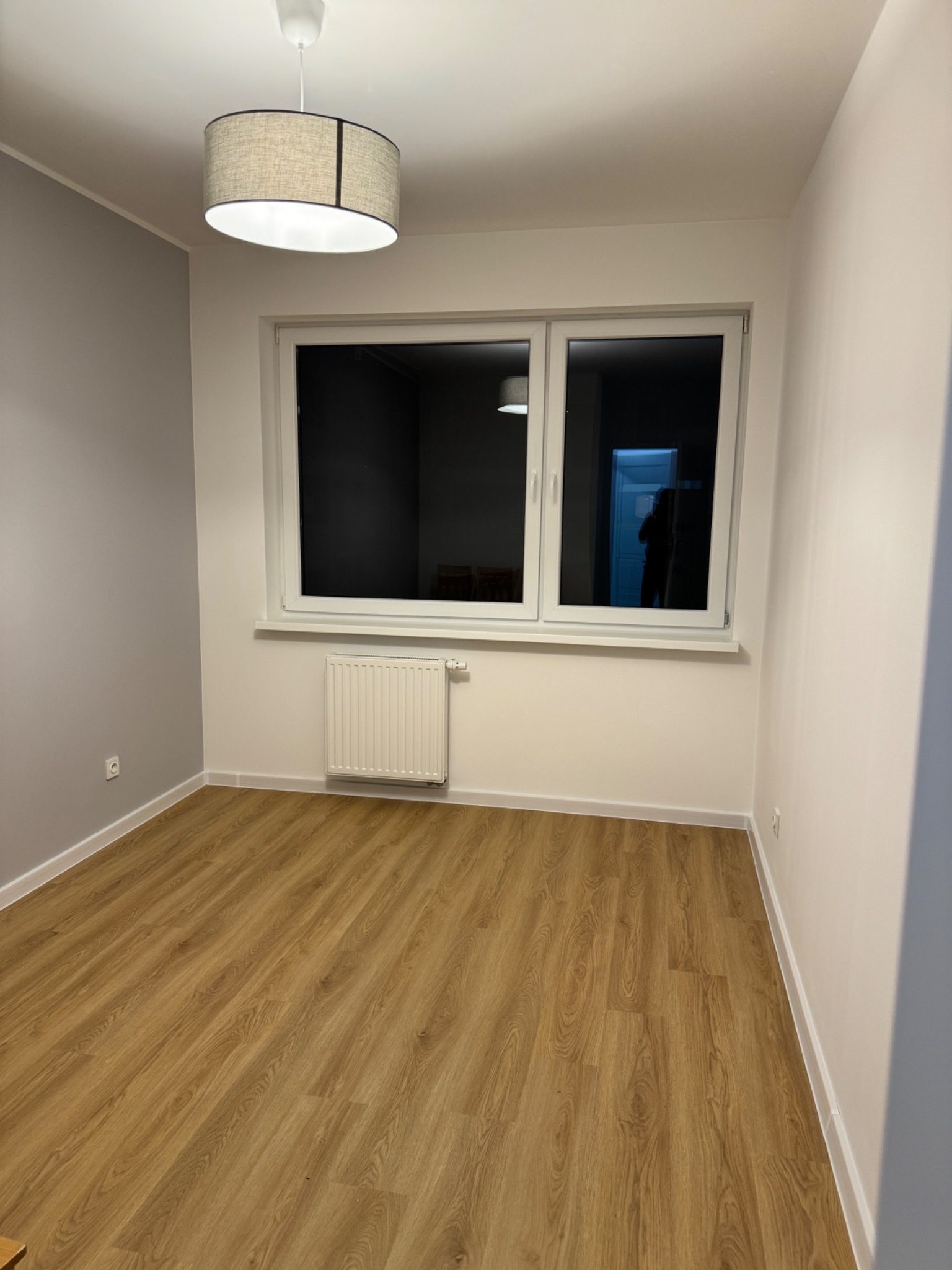 Pusty pokój po remoncie z panelami podłogowymi w odcieniach brązu, świeżo pomalowanymi ścianami w kolorach bieli i szarości, grzejnikiem pod oknem i nowoczesną lampą sufitową.