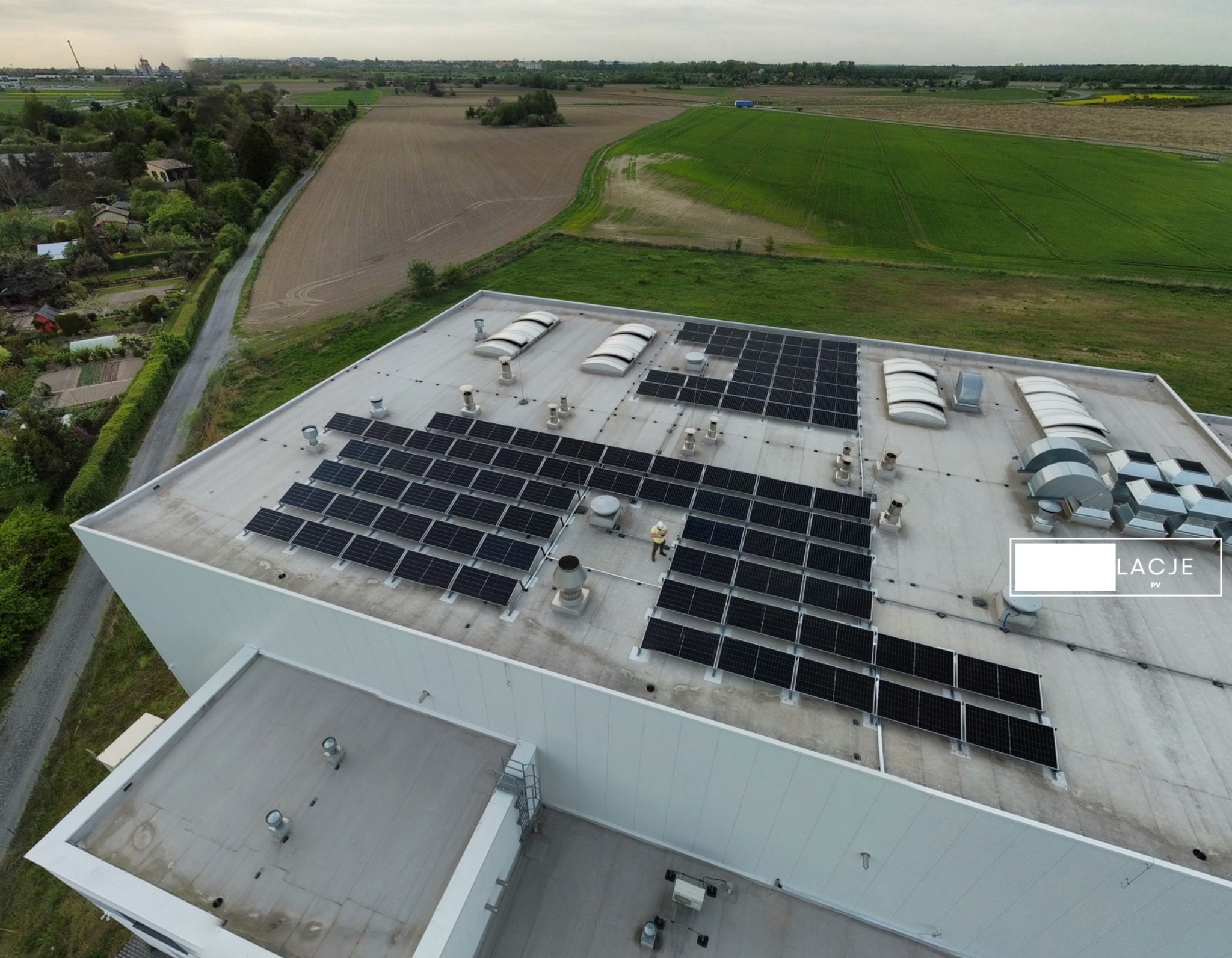 Widok z drona na dach budynku przemysłowego z zainstalowanymi panelami słonecznymi; w tle pola uprawne i drzewa, na dachu widoczny pracownik w kasku.