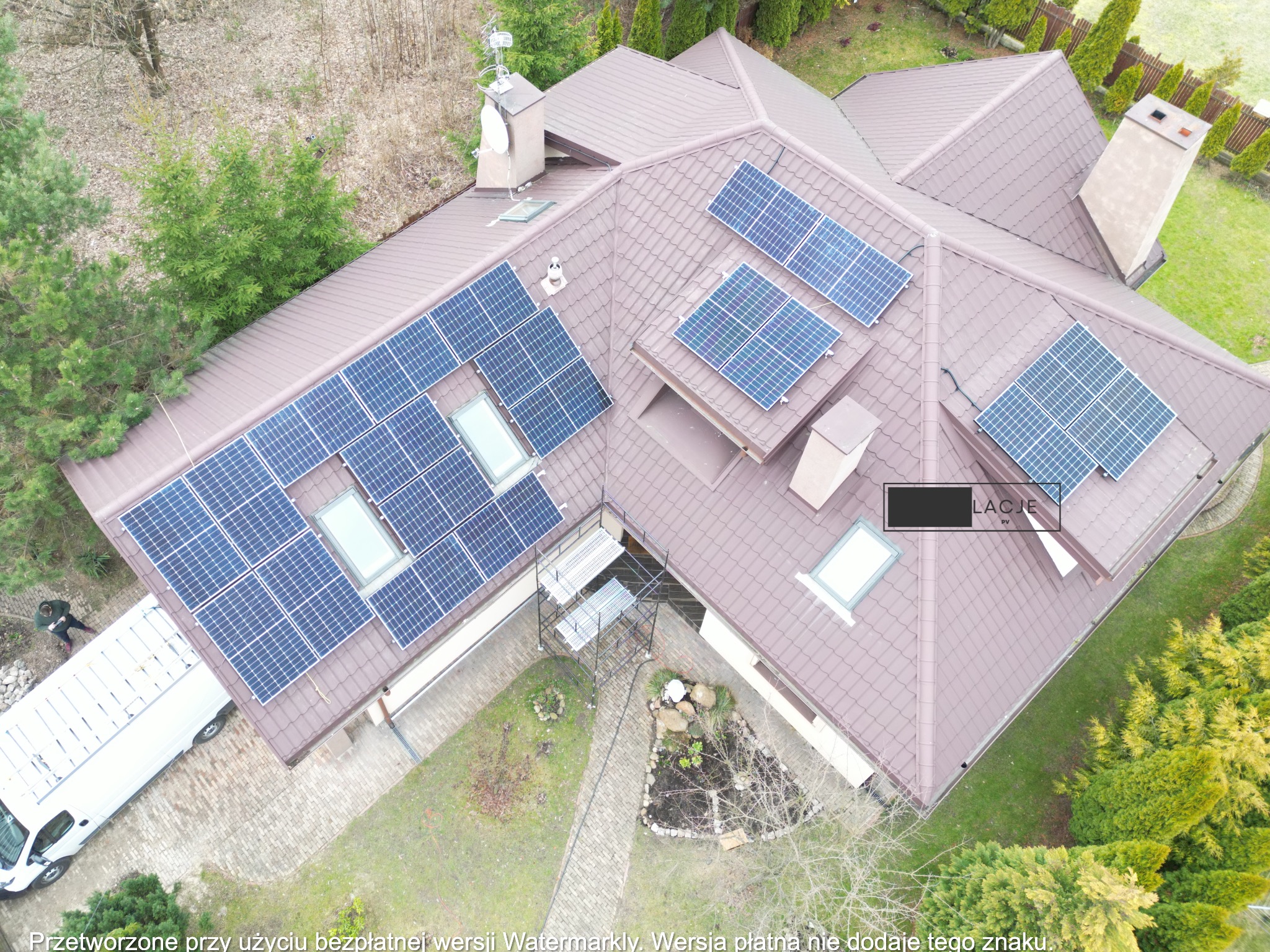 Widok z drona na dom z brązowym dachem, na którym zamontowane są panele słoneczne; obok zaparkowany biały samochód dostawczy, a w tle drzewa.