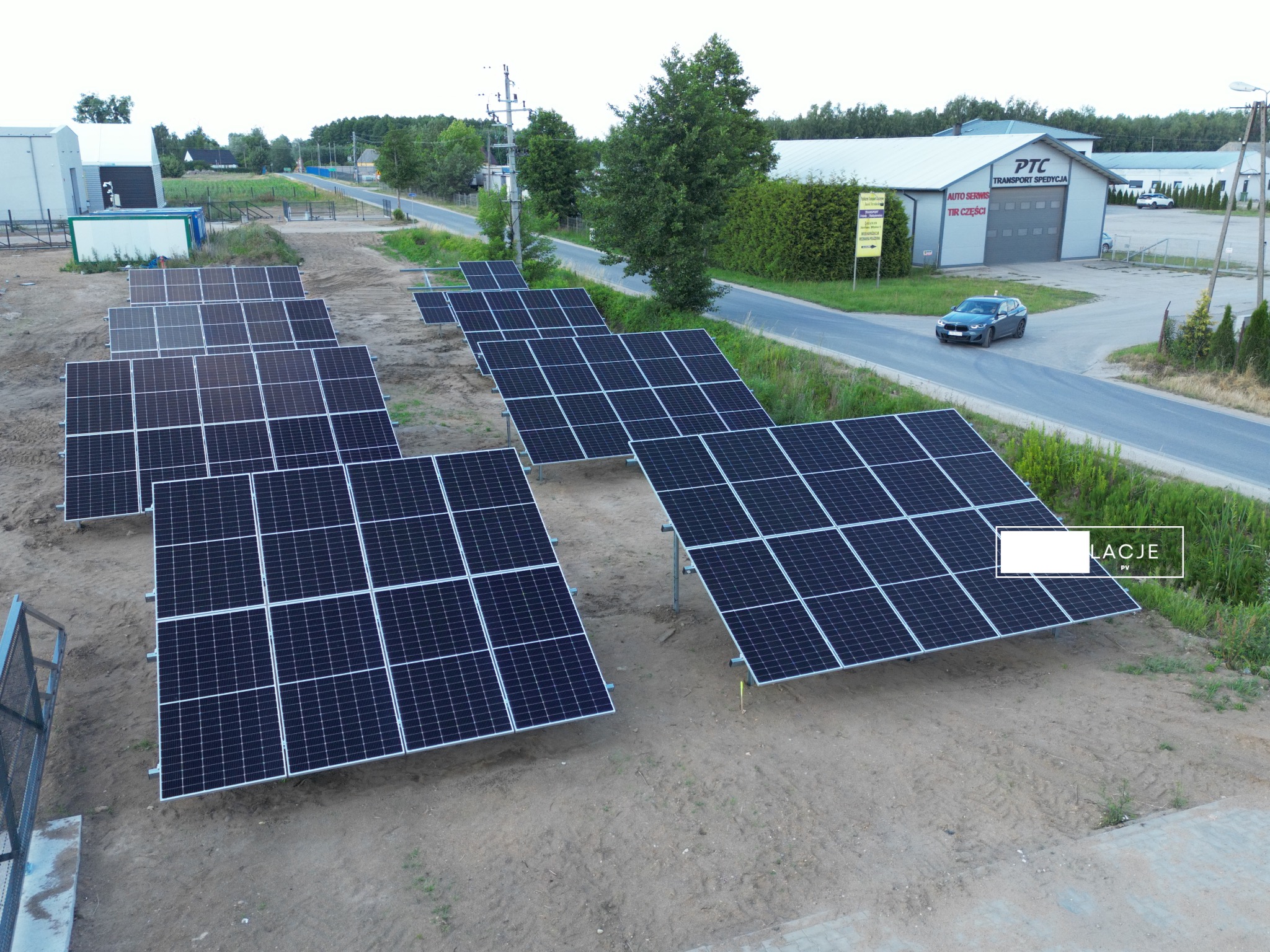 Rozległa instalacja fotowoltaiczna na gruncie, widok z lotu ptaka, z budynkiem PTC Transport Spedycja w tle i przejeżdżającym samochodem.