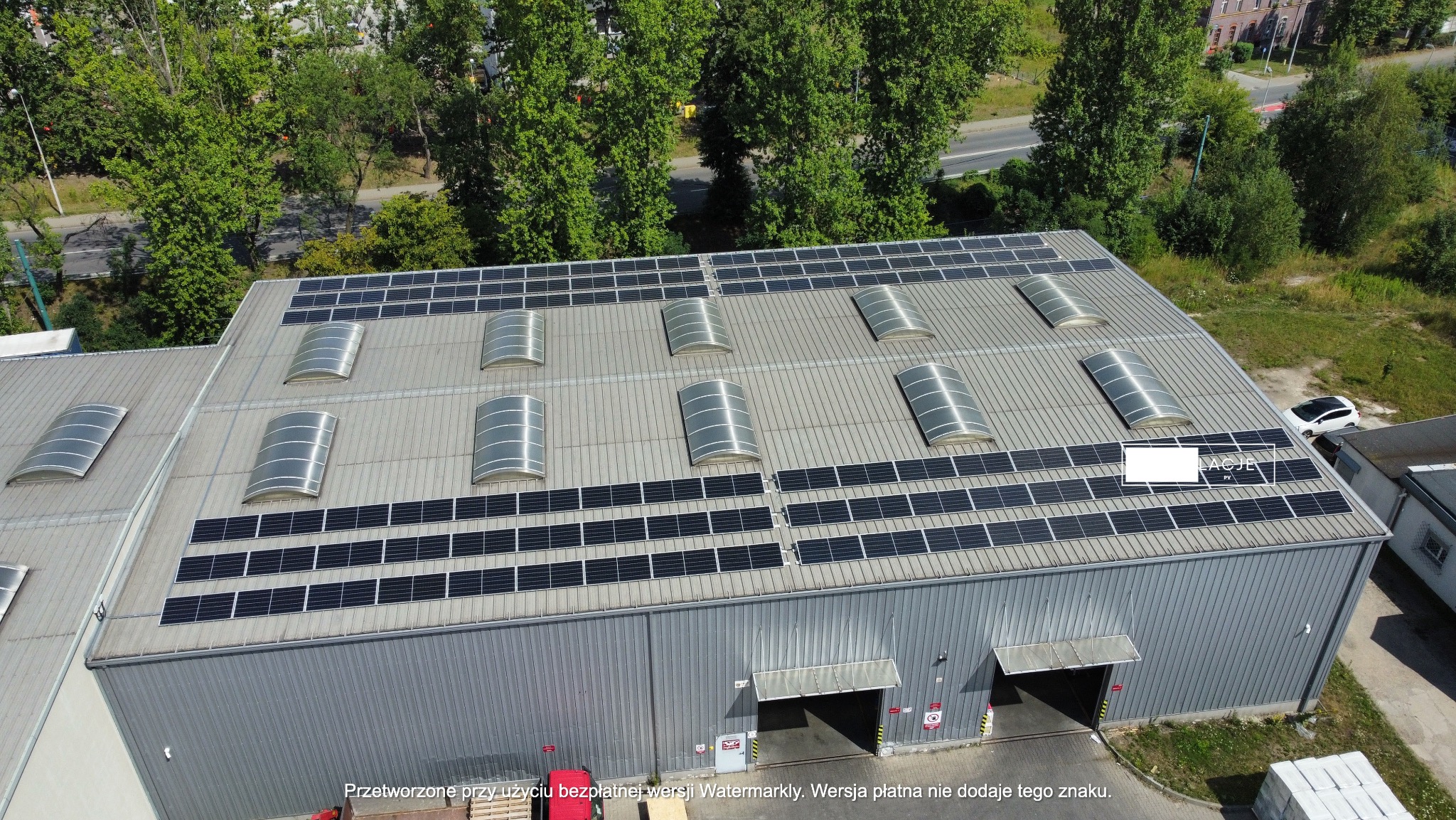 Widok z góry na dużą halę przemysłową z szarymi panelami słonecznymi i świetlikami na dachu, otoczoną zielenią drzew i ulicą w tle. Dwa otwarte wjazdy do hali.