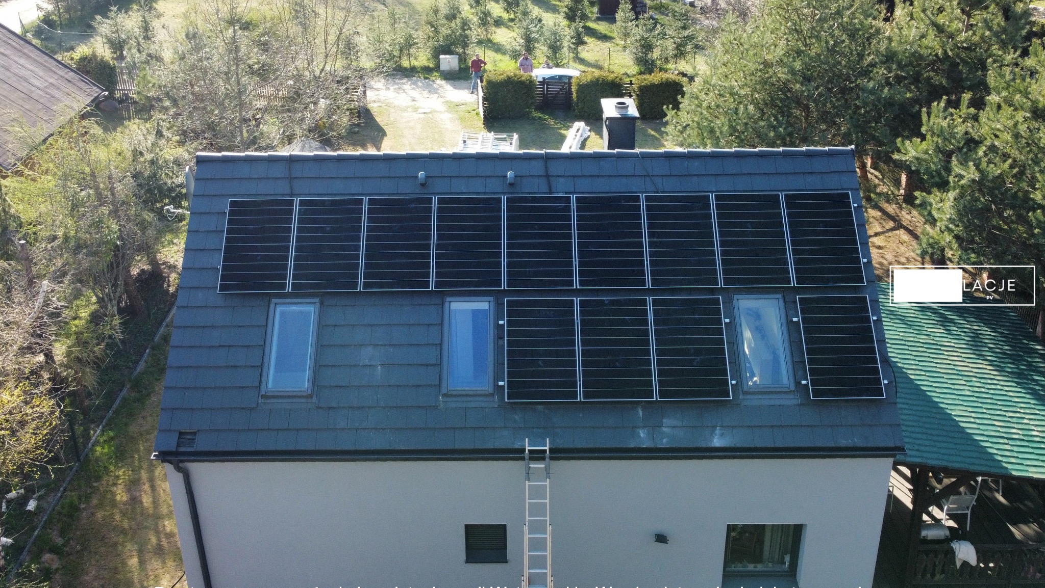 Instalacja paneli słonecznych na dachu domu jednorodzinnego z oknami dachowymi, widok z góry, drabina oparta o ścianę, dwóch mężczyzn w oddali na trawniku.