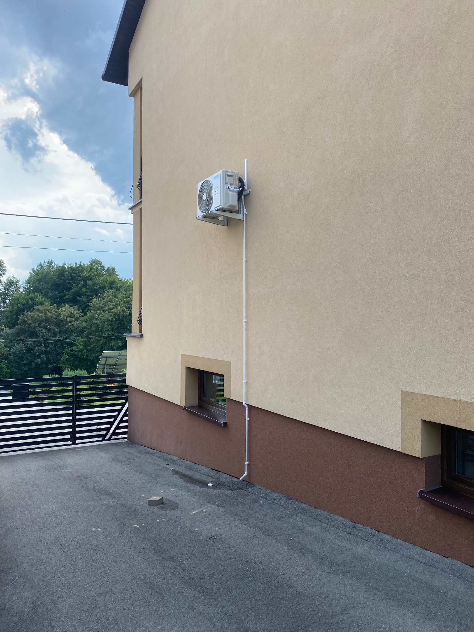 Zewnętrzna jednostka klimatyzacji zamontowana na elewacji budynku z beżowym tynkiem, widoczne rury odprowadzające skropliny, brama w tle.