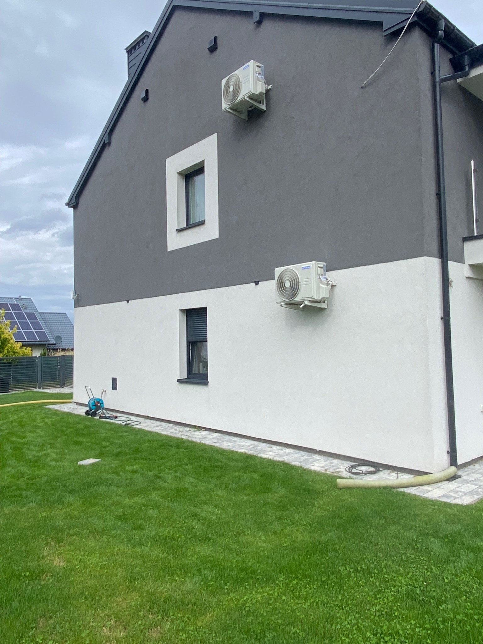 Biało-szary dom z dwoma zamontowanymi klimatyzatorami na elewacji. Na trawniku wąż ogrodowy i wózek do podlewania. Widoczne panele słoneczne na dachu sąsiedniego budynku.