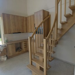 Graf Bud Mykola Yuskevych - Jasne, drewniane schody w nowoczesnym wnętrzu z zabudową kuchenną w tle. Widoczne duże okno i częściowo rozpakowane materiały budowlane.