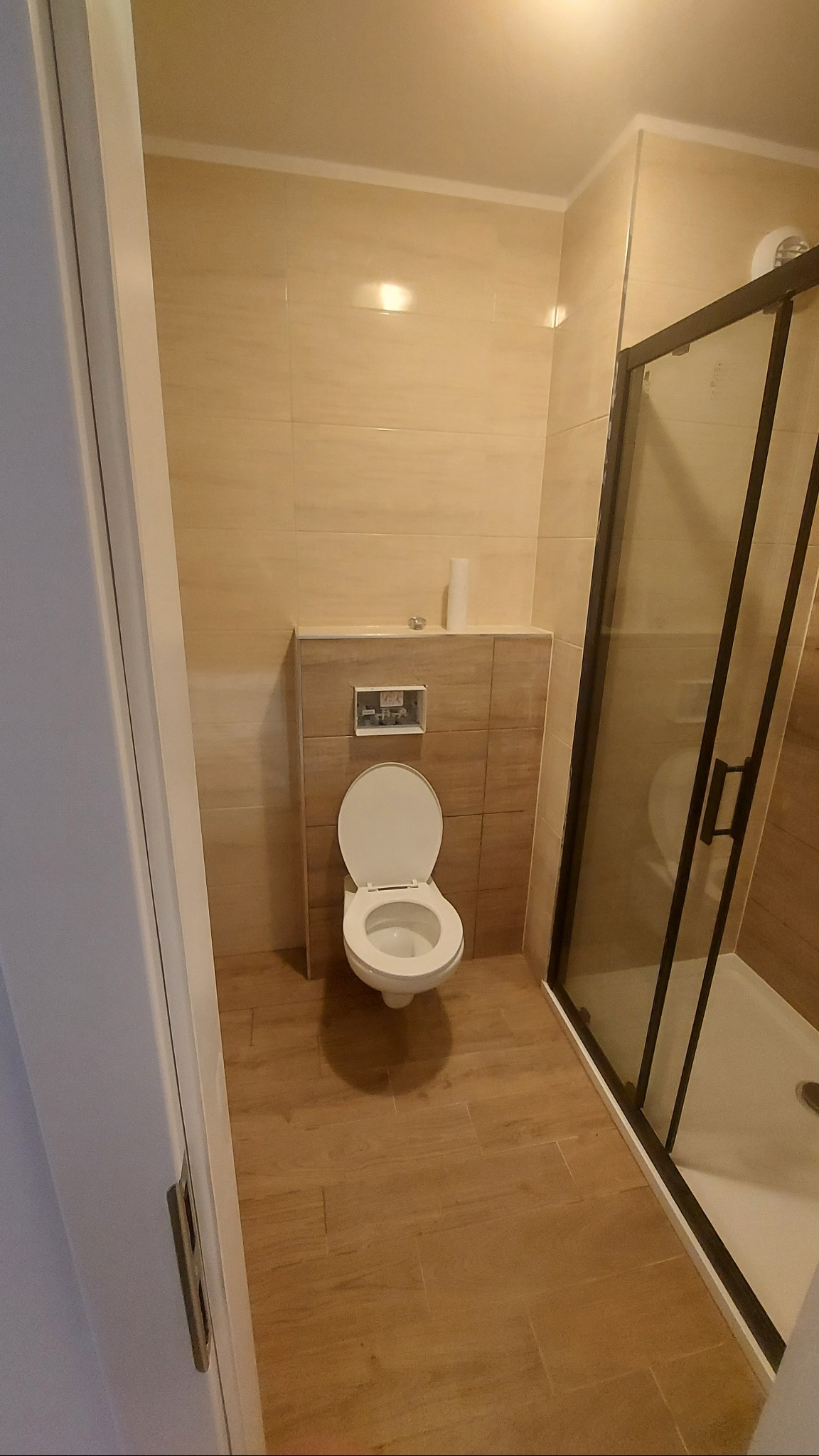 Wykończona łazienka z beżowymi płytkami, kabiną prysznicową z czarną ramą i toaletą. Widok z góry na całe pomieszczenie, ukazujący estetyczne detale i aranżację.