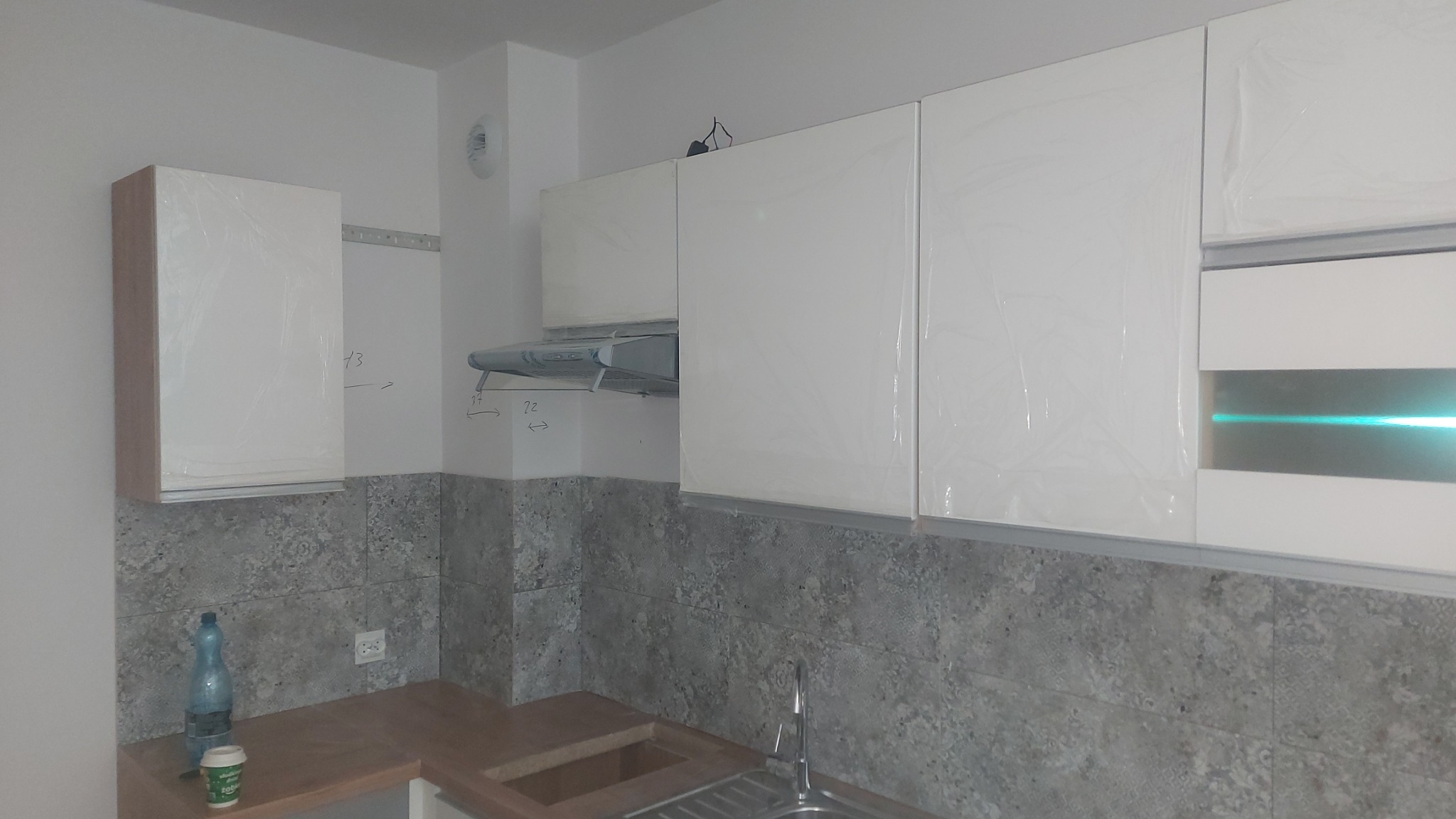 Nowoczesna kuchnia w trakcie montażu: białe szafki z folią ochronną, szary wzór płytek na ścianie, widoczne przyłącza i notatki na ścianie.