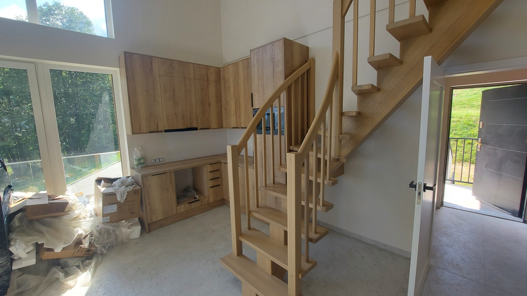 Jasne, drewniane schody w nowoczesnym wnętrzu z zabudową kuchenną w tle. Widoczne duże okno i częściowo rozpakowane materiały budowlane.