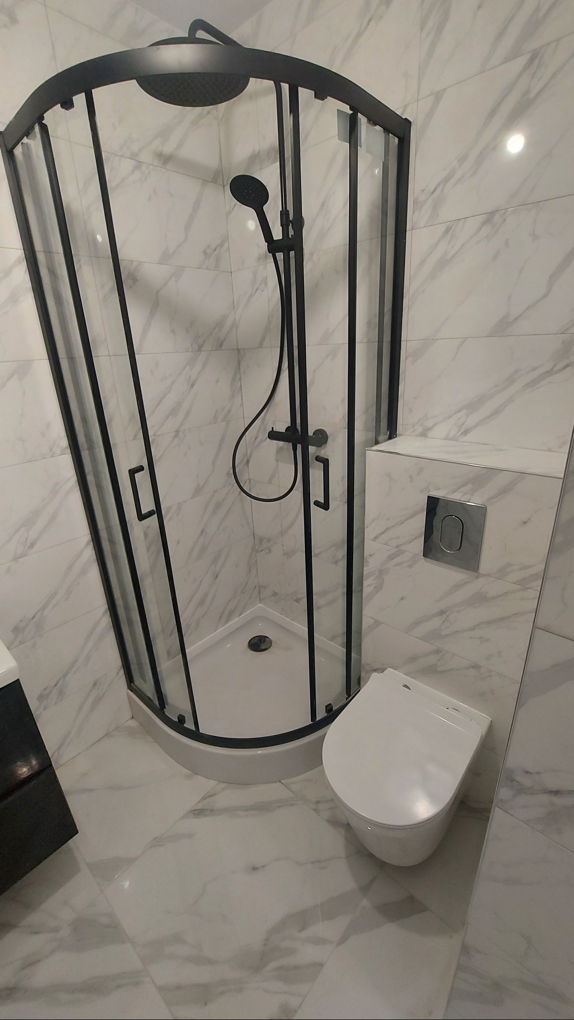 Nowoczesna kabina prysznicowa z czarnymi profilami i deszczownicą, biała toaleta wisząca, marmurowe płytki na ścianach i podłodze. Minimalistyczny design łazienki.