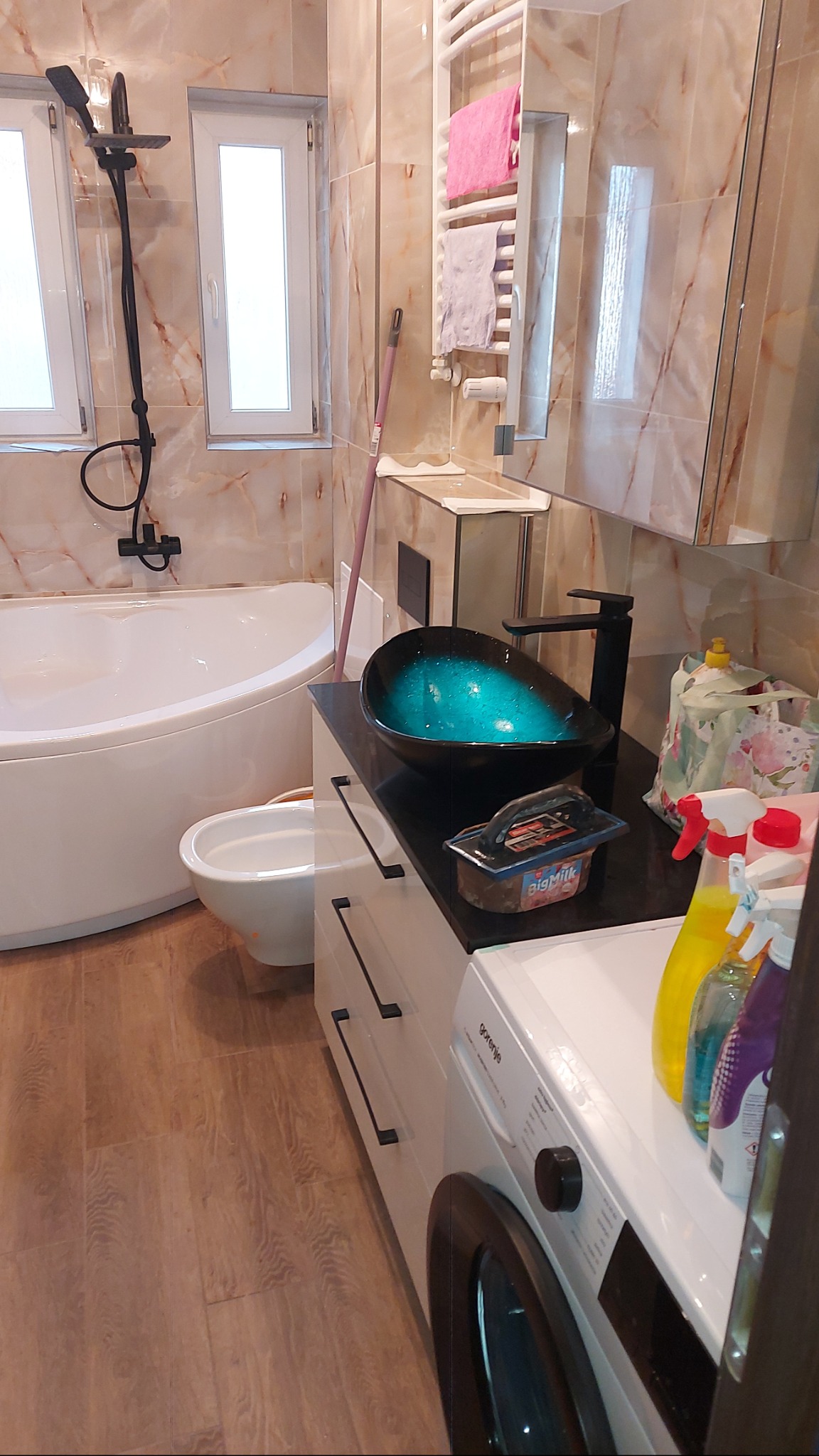 Wykończona łazienka z umywalką w kształcie misy, białą wanną narożną i pralką. Na blacie narzędzia glazurnicze. Płytki imitujące marmur na ścianach.