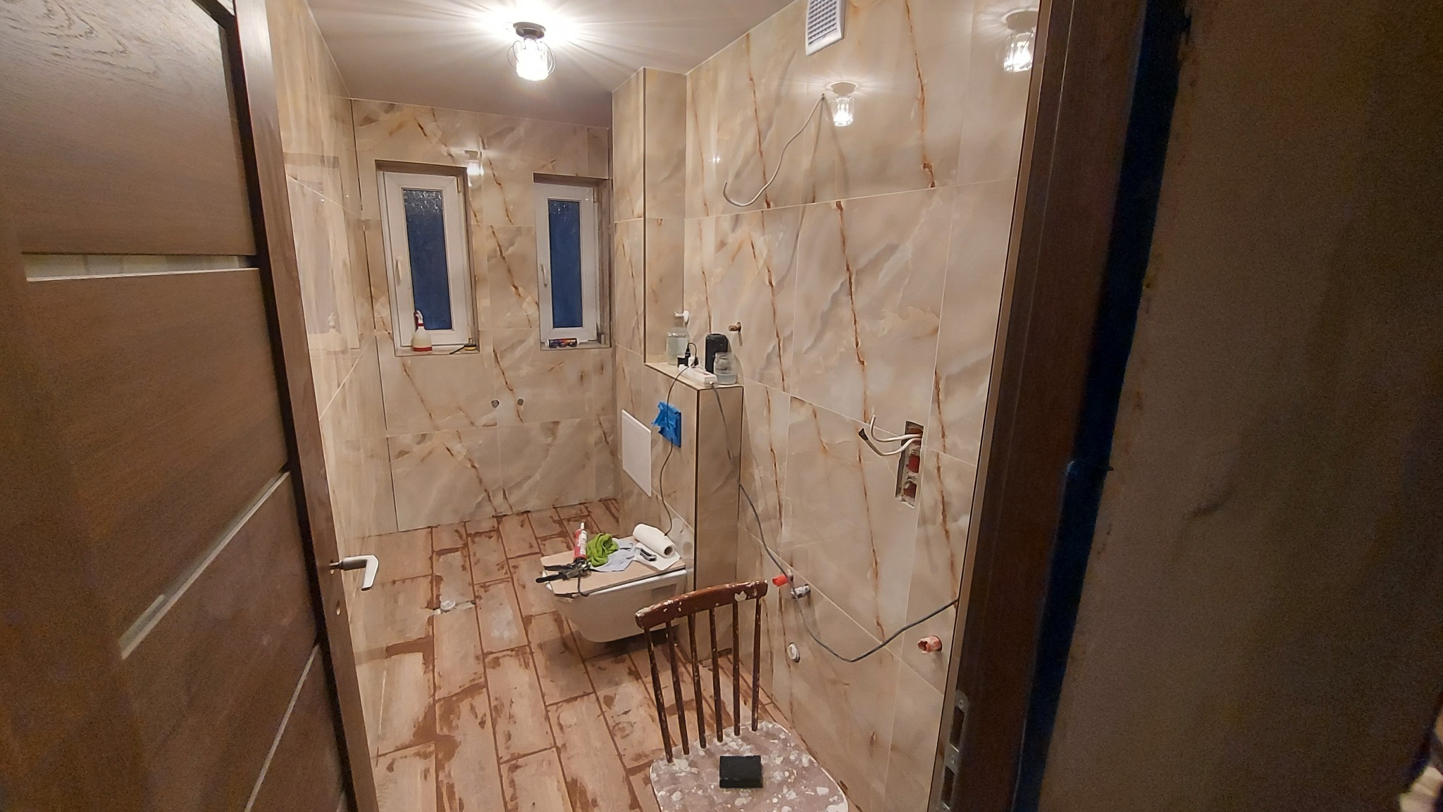 Remont łazienki w trakcie: beżowe płytki imitujące marmur na ścianach, drewnopodobne płytki na podłodze, częściowo zamontowana toaleta, widoczne przewody elektryczne.