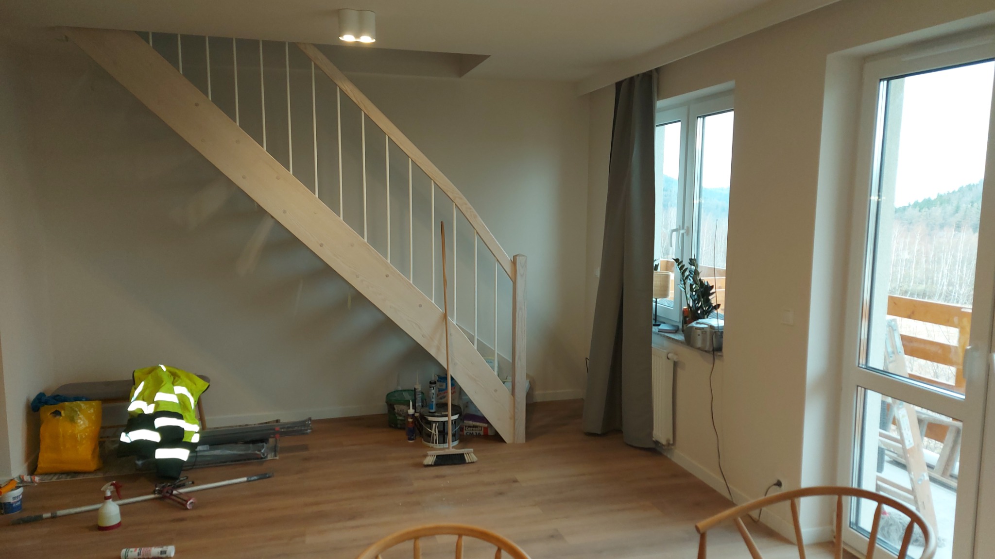 Jasne drewniane schody w nowym wnętrzu, widoczne narzędzia i odzież robocza, okno z widokiem na zewnątrz. Podłoga z paneli, neutralna kolorystyka ścian.