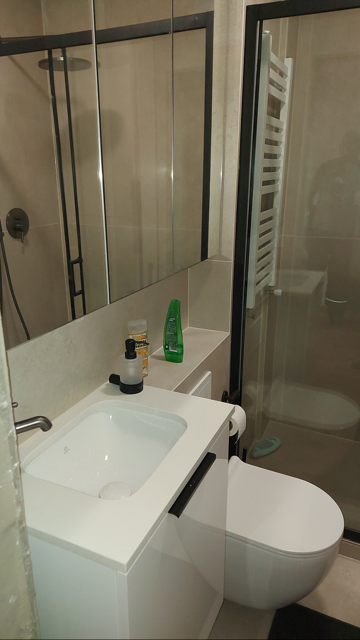 Nowoczesna łazienka z białą umywalką nablatową, toaletą i kabiną prysznicową z czarnymi ramami. Minimalistyczny design, beżowe płytki, oświetlenie punktowe.