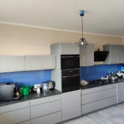 Jasnoszara kuchnia z niebieskim panelem nad blatem, widoczne sprzęty AGD, w tle dwie lampy wiszące z czarnym kloszem w formie klatki.