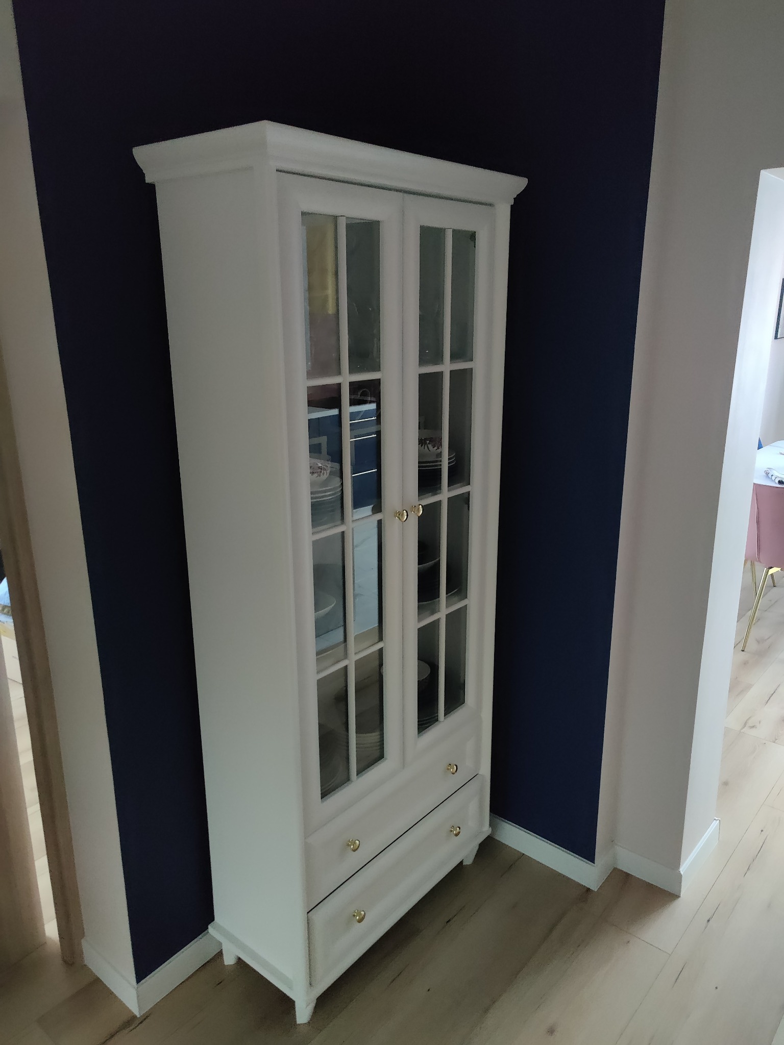 Biały kredens z przeszklonymi drzwiami i dwoma szufladami, ustawiony na tle ściany pomalowanej na granatowo, widoczny fragment parkietu i jasnej ściany w sąsiednim pomieszczeniu.