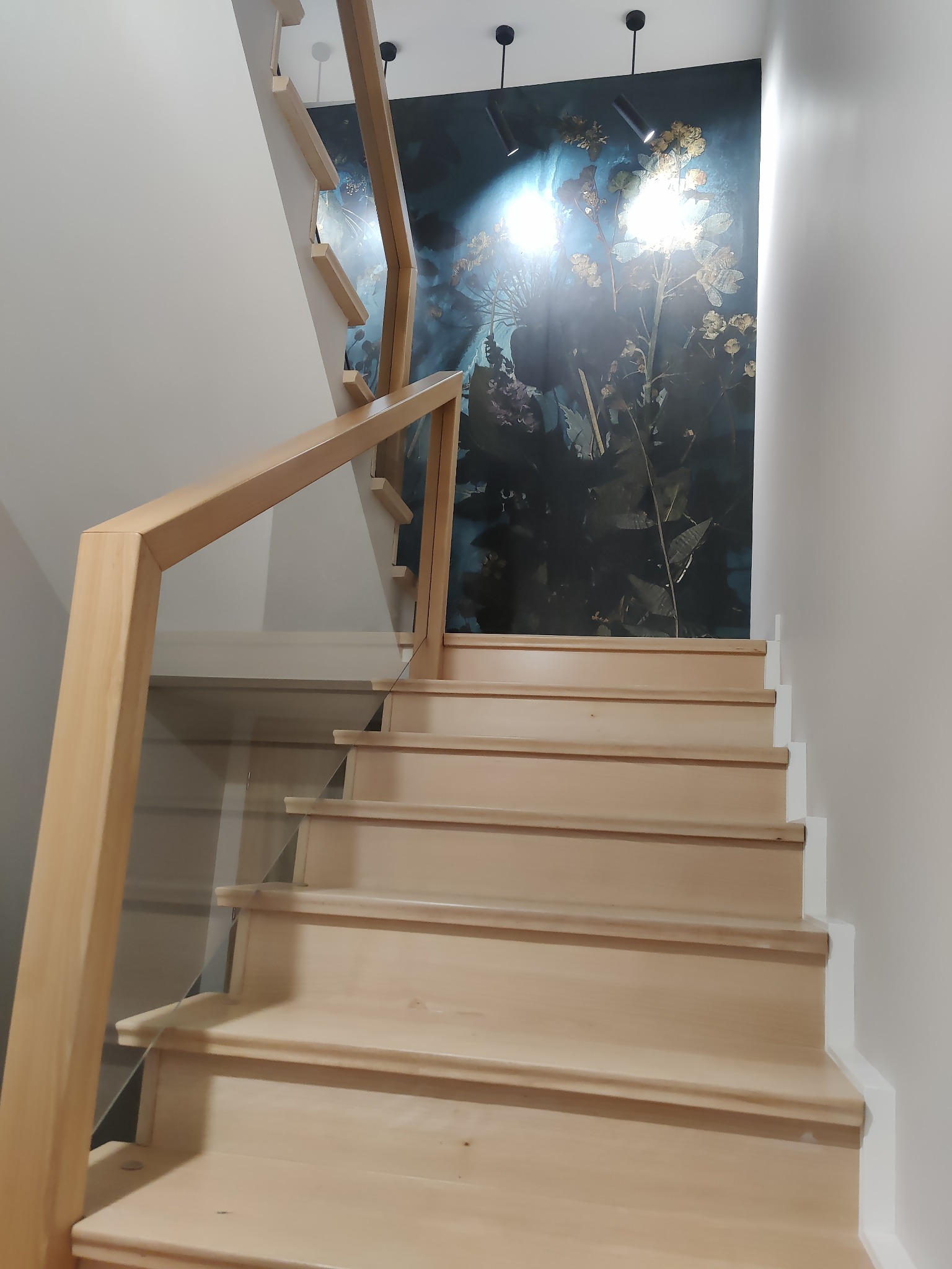 Jasne, drewniane schody z przeszkloną balustradą prowadzące na górę, z ciemną tapetą w kwiaty w tle i punktowym oświetleniem sufitowym.