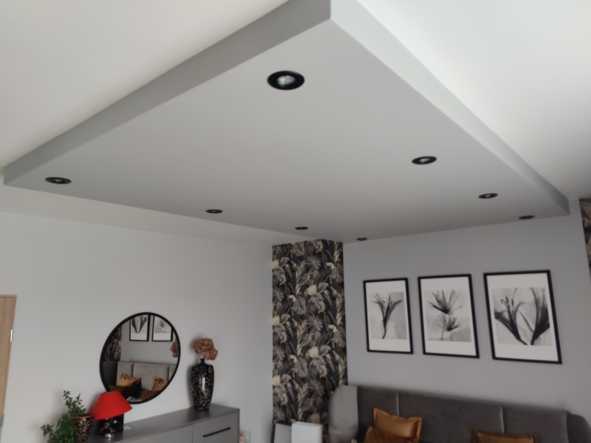 Podwieszany sufit z oświetleniem punktowym w salonie, dekoracje ścienne, tapeta w roślinne wzory, szara komoda z lustrem i wazonem.