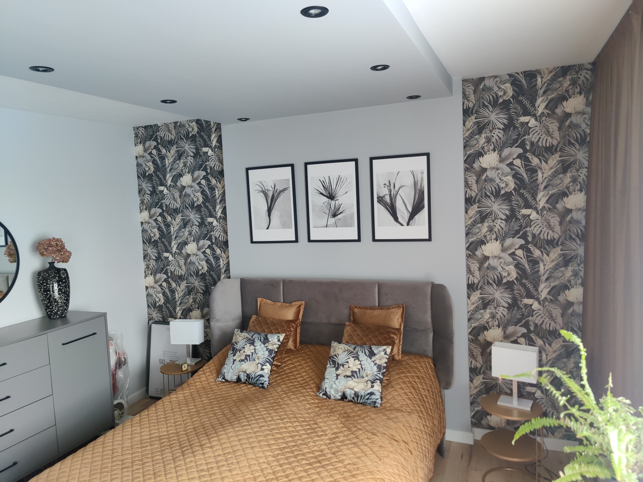 Sypialnia w odcieniach szarości i złota z tapetą w motywy roślinne, trzema obrazami w czarnych ramkach i rośliną doniczkową.