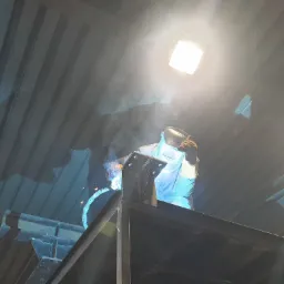 Pracownik w masce spawalniczej łączy elementy konstrukcji stalowej metodą spawania elektrycznego w hali produkcyjnej, widoczny dym i iskry.