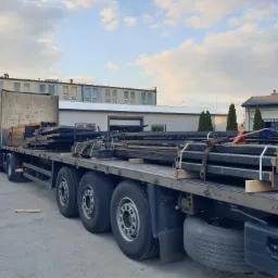 Transport konstrukcji stalowych na naczepie ciężarówki, zabezpieczonych pasami, z widocznymi elementami z otworami montażowymi, na tle budynków przemysłowych i błękitnego nieba z chmurami.