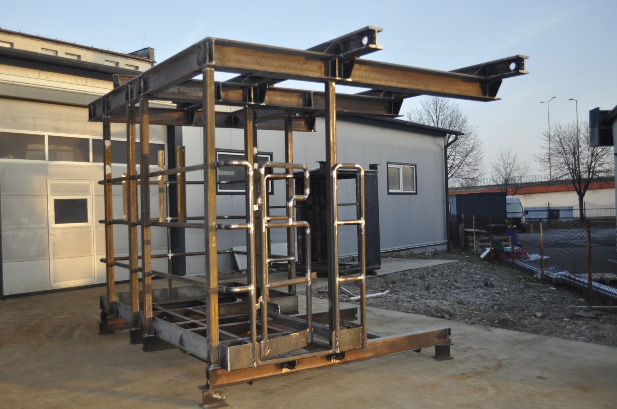 Szkielet stalowej konstrukcji hali w trakcie budowy, widoczne elementy spawane i przygotowane do montażu, tło stanowi budynek biurowy i plac budowy.