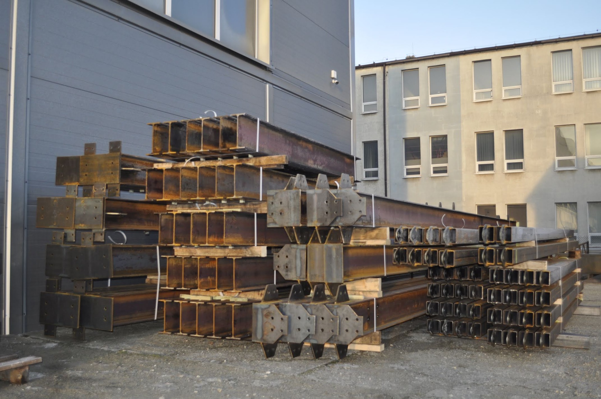 Stalowe belki konstrukcyjne, z widocznymi elementami mocującymi i otworami, ułożone w stosy na placu budowy, z budynkami w tle.