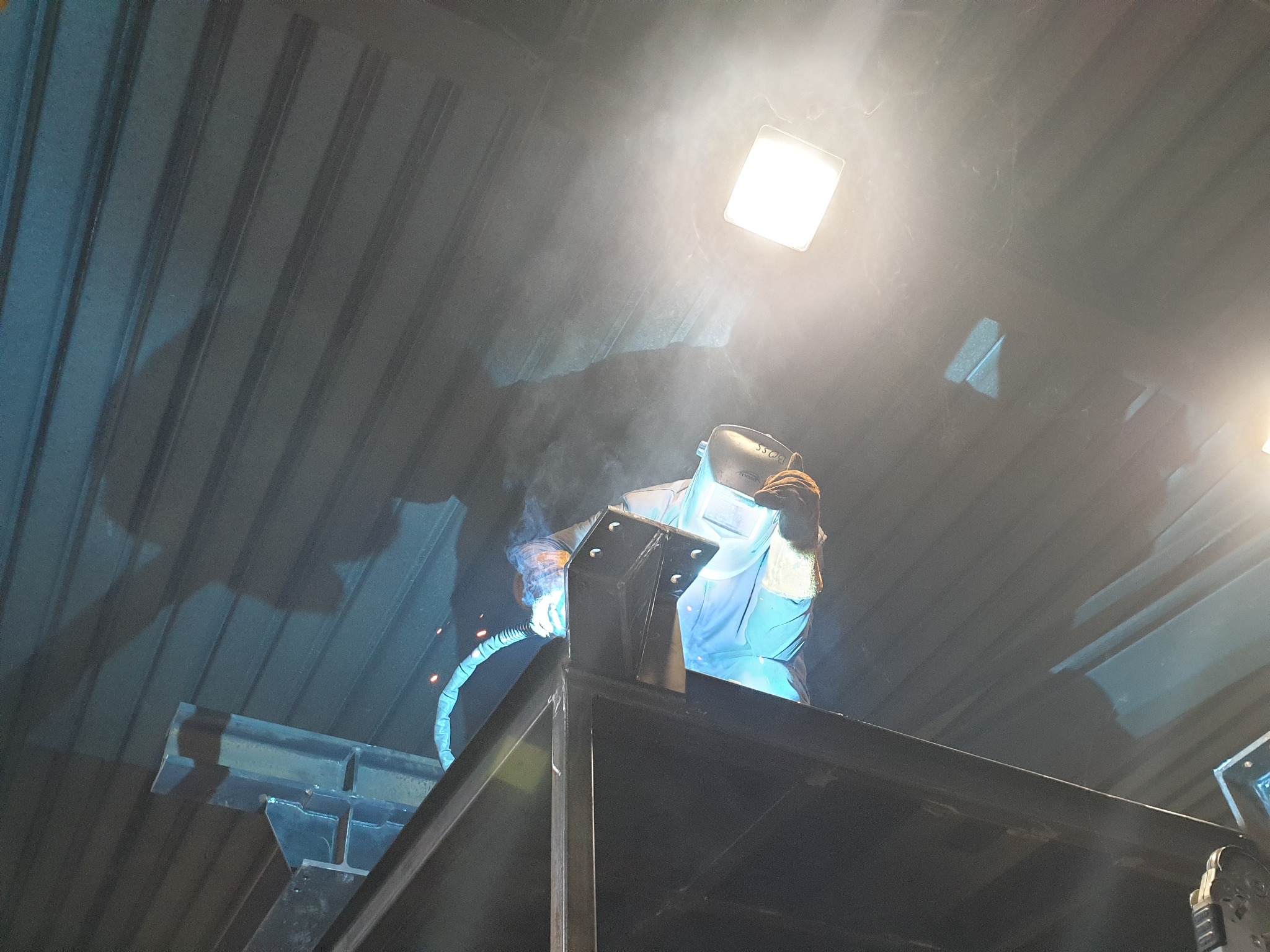 Pracownik w masce spawalniczej łączy elementy konstrukcji stalowej metodą spawania elektrycznego w hali produkcyjnej, widoczny dym i iskry.