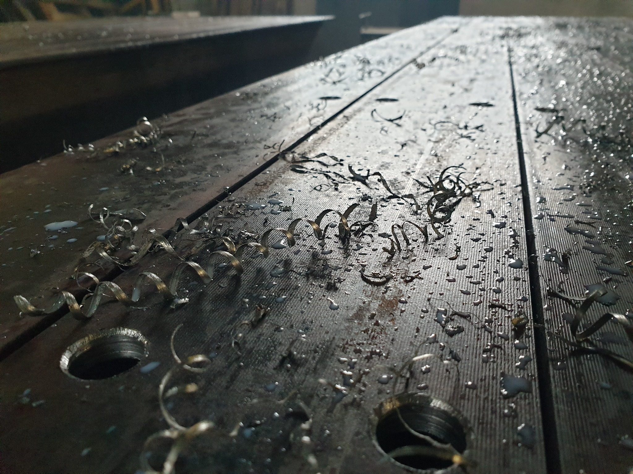 Metalowa płyta z wywierconymi otworami, pokryta wiórami i kroplami chłodziwa, oświetlona z góry, widoczne ślady obróbki skrawaniem.