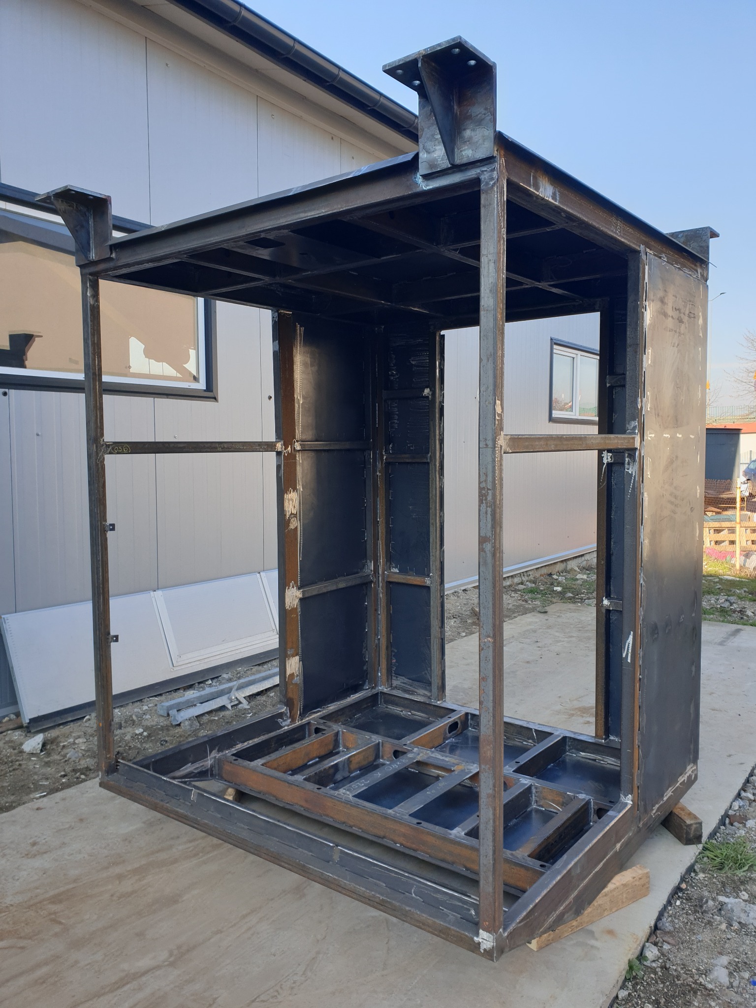 Surowa, spawana konstrukcja stalowa hali w trakcie budowy, widoczna na zewnątrz, z elementami mocującymi na dachu i częściowym wypełnieniem ścian, umieszczona na betonowej podstawie.