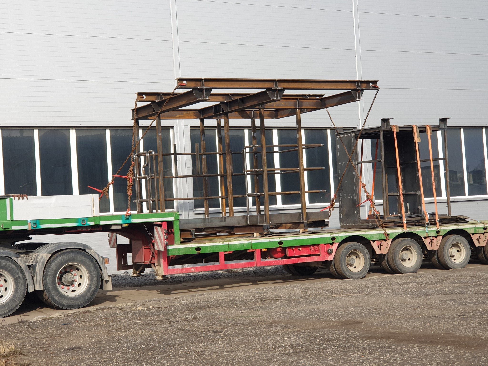 Transport konstrukcji stalowej na naczepie niskopodwoziowej, zabezpieczonej łańcuchami i pasami transportowymi, na tle hali z oknami.