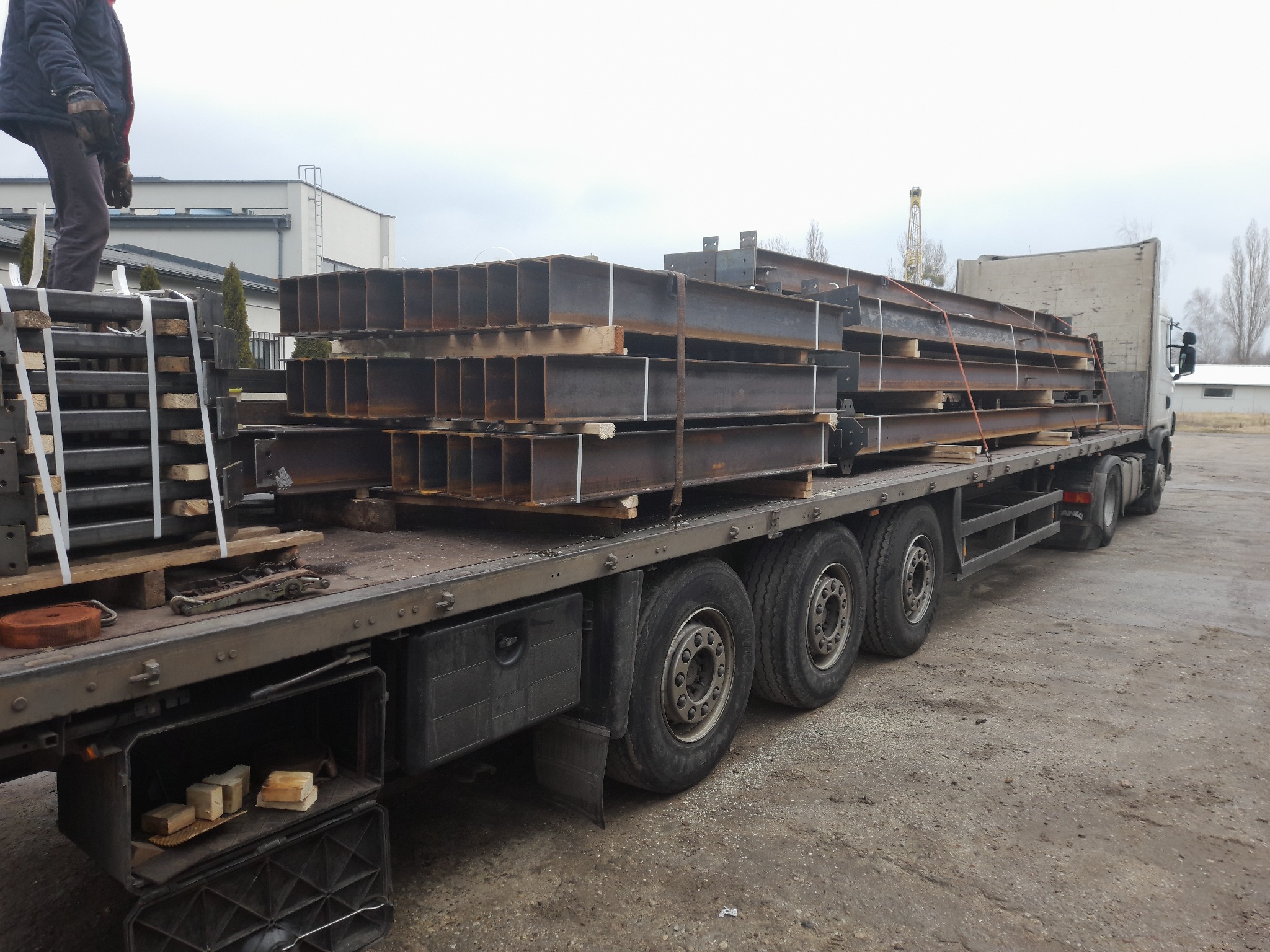 Transport belek stalowych na naczepie ciężarówki, zabezpieczonych pasami, z widocznym fragmentem osoby na platformie załadunkowej.