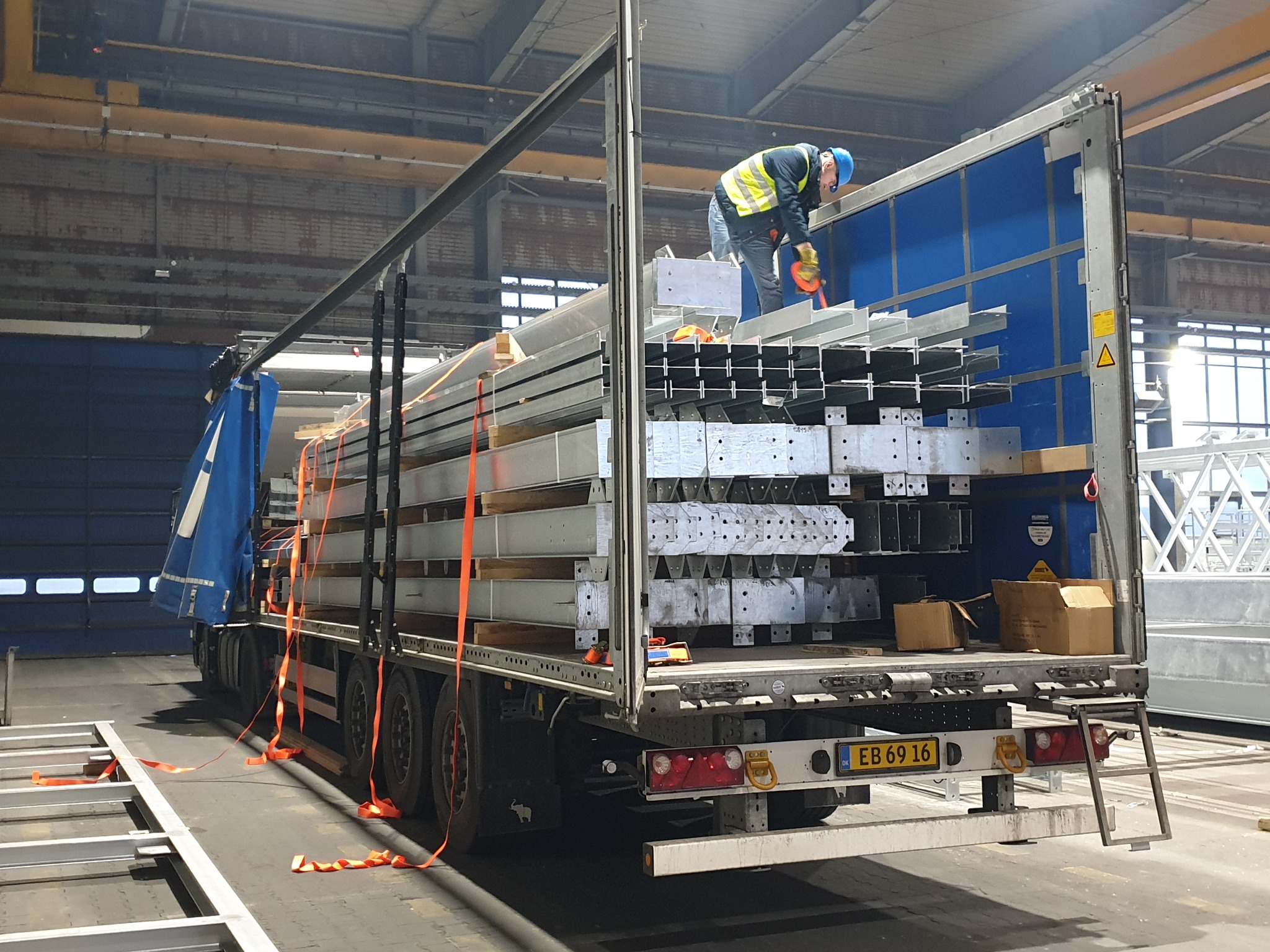 Pracownik w kasku i kamizelce odblaskowej zabezpiecza ładunek konstrukcji stalowych na naczepie ciężarówki, widoczne elementy mocujące i kartony.