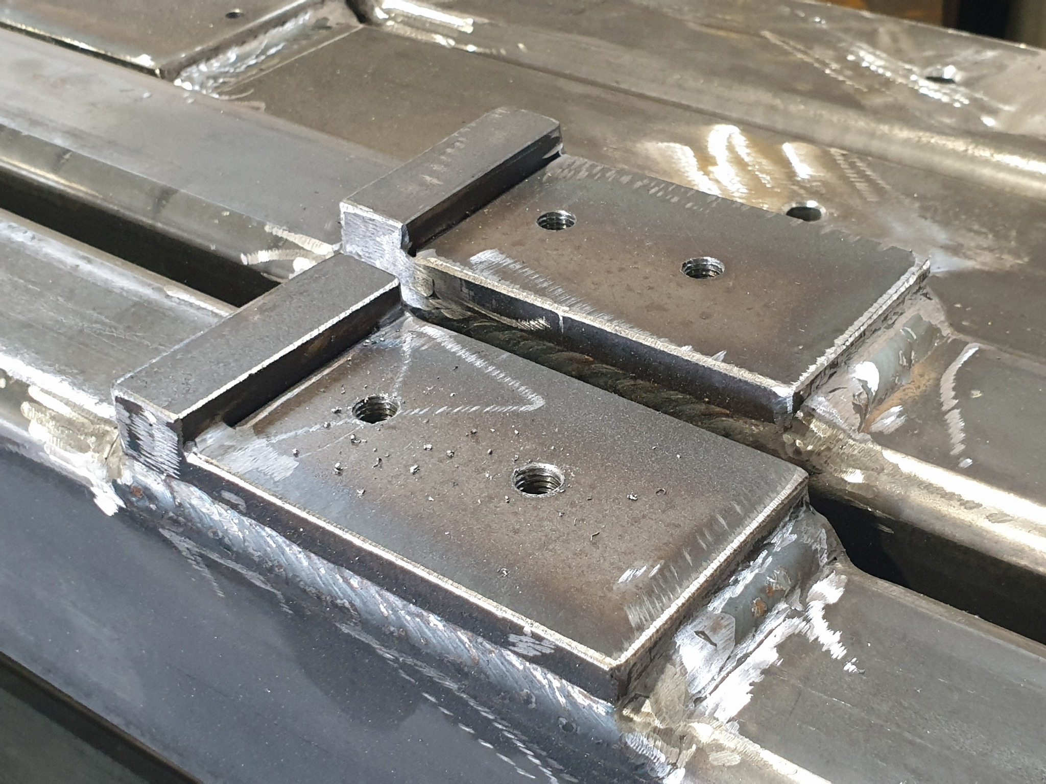 Metalowe elementy połączone spawami, widoczne ślady obróbki i otwory gwintowane, detale konstrukcji stalowej z bliska.