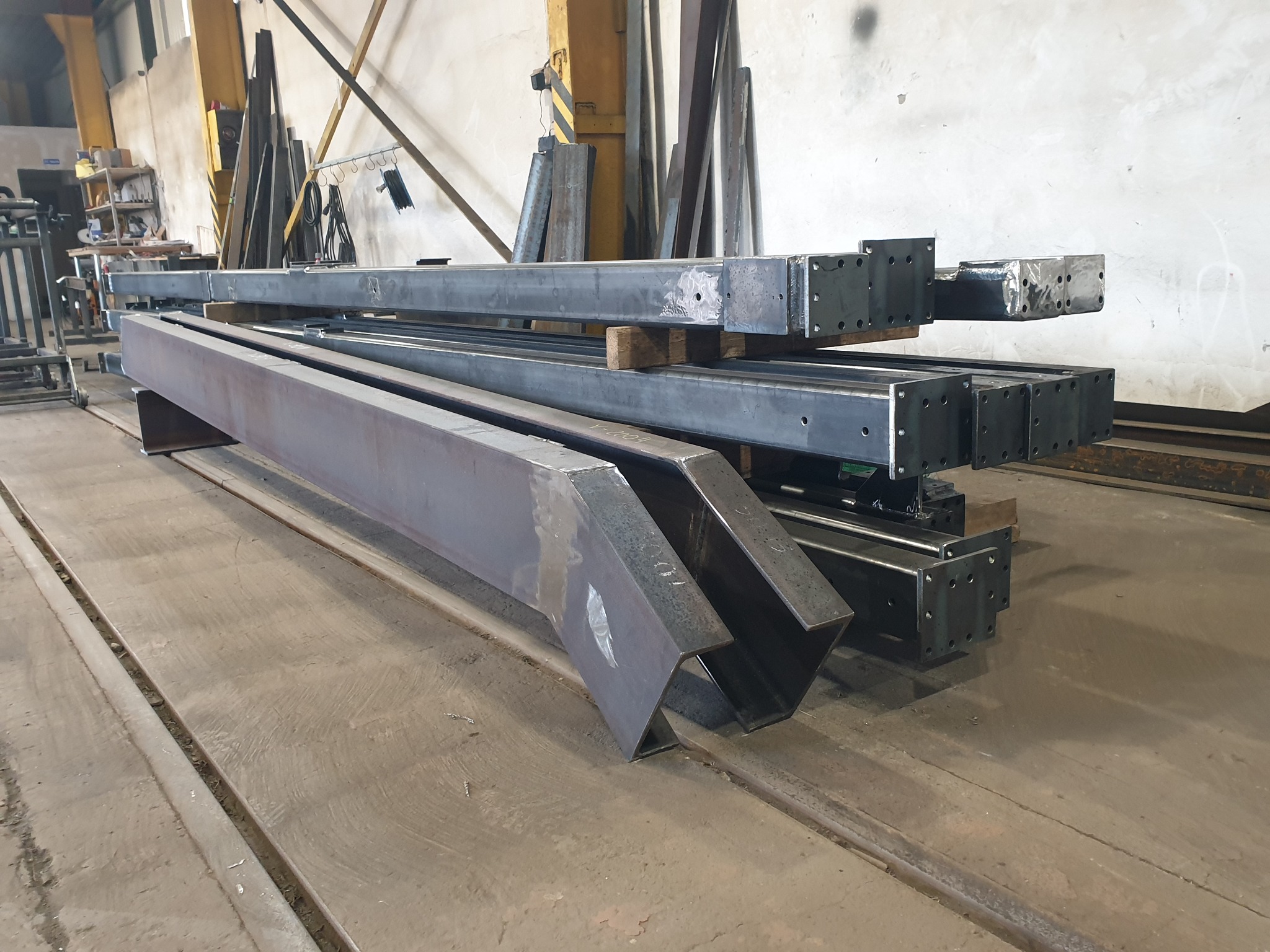 Stalowe elementy konstrukcyjne, belki i słupy, ułożone na drewnianych podkładkach w hali produkcyjnej, widoczne ślady po spawaniu.