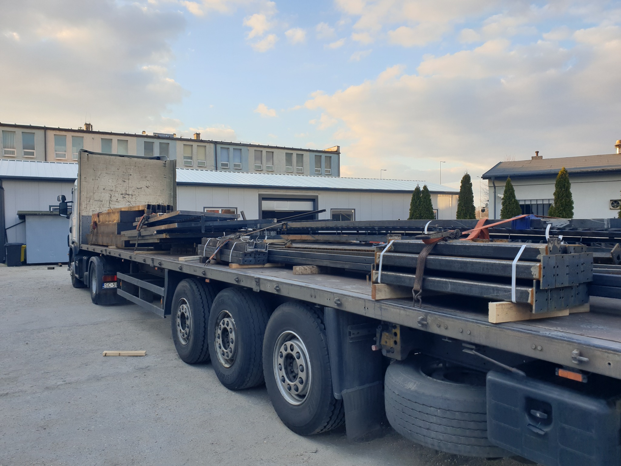 Transport konstrukcji stalowych na naczepie ciężarówki, zabezpieczonych pasami, z widocznymi elementami z otworami montażowymi, na tle budynków przemysłowych i błękitnego nieba z chmurami.