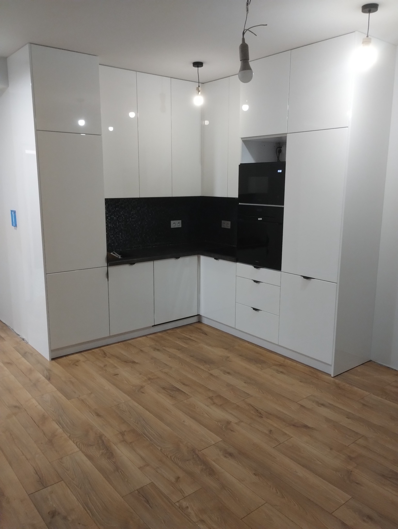 Nowoczesna kuchnia z białymi, lakierowanymi frontami, czarnym panelem nad blatem i zabudowanymi urządzeniami AGD, oświetlona trzema lampami wiszącymi z odsłoniętymi żarówkami.