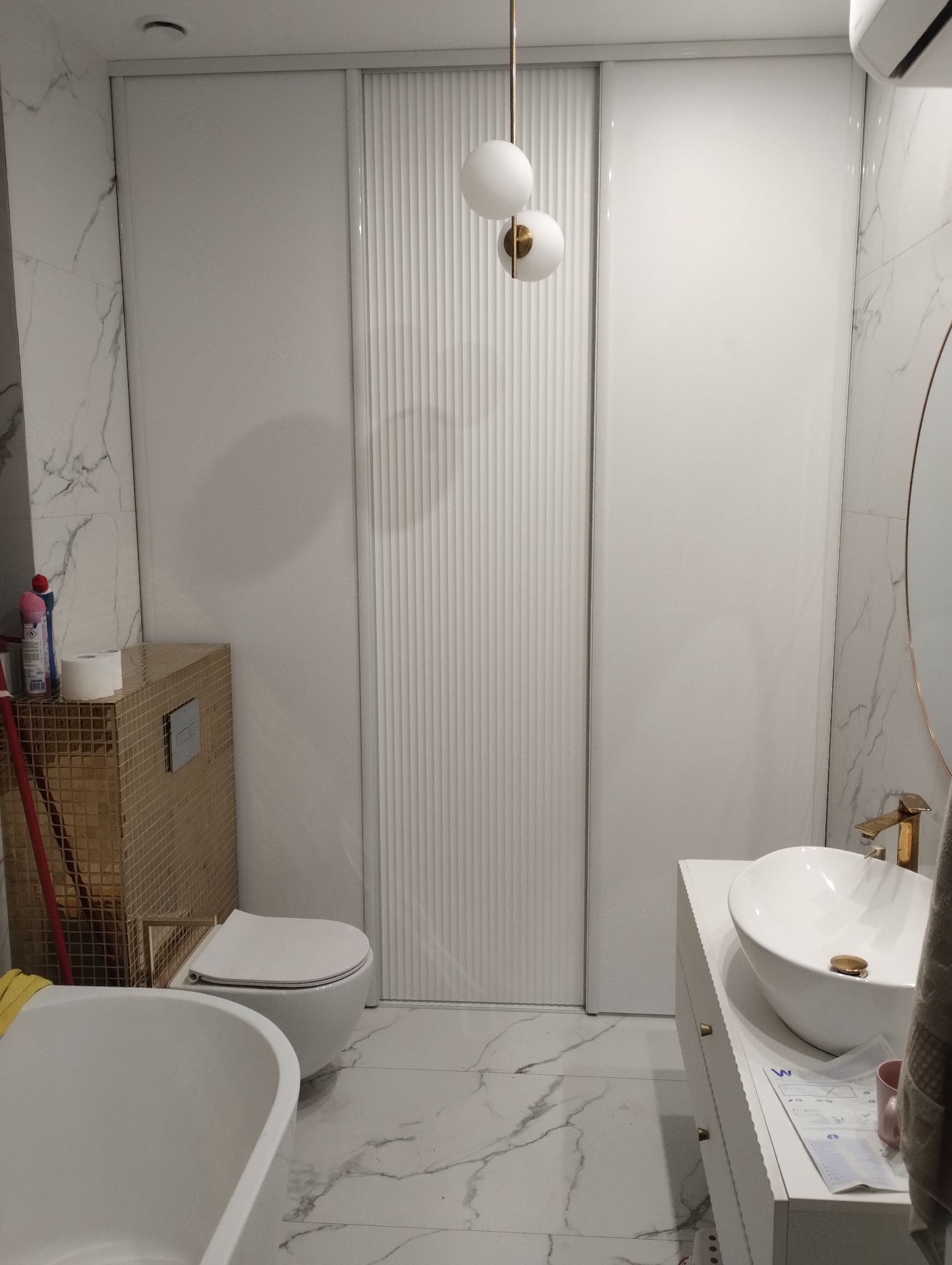 Nowoczesna łazienka z marmurowymi płytkami, złotymi akcentami i przesuwnymi drzwiami z pionowymi żłobieniami, oświetlona wiszącą lampą z białymi kulami.