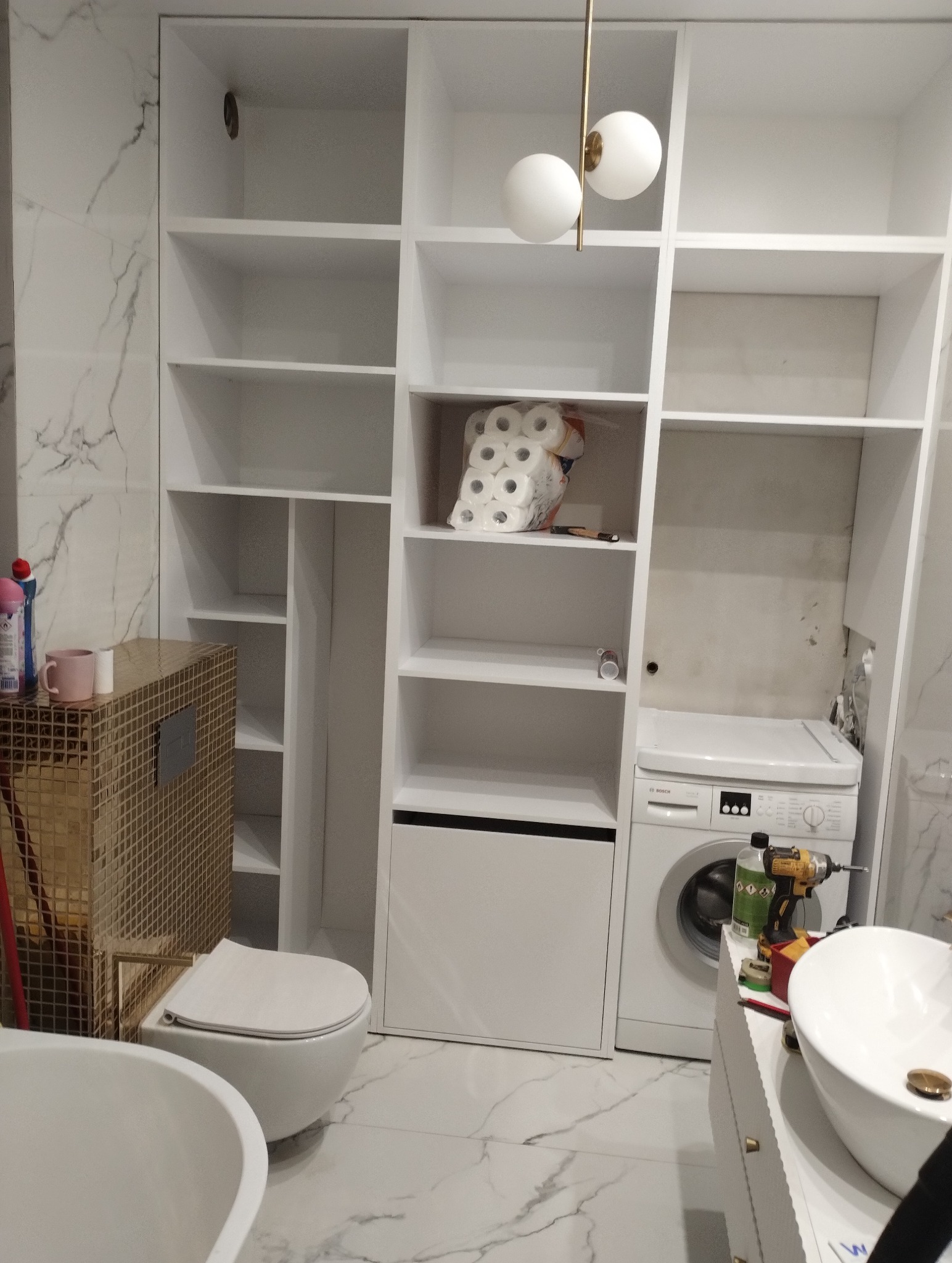 Biała zabudowa meblowa w łazience z pralką, toaletą i umywalką na szafce, w trakcie montażu widoczna wiertarka i opakowanie papieru toaletowego.