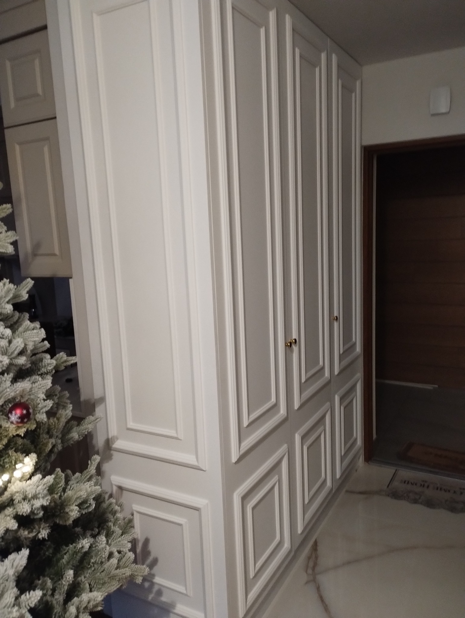 Biała szafa wnękowa z dekoracyjnymi frezowaniami, złotymi uchwytami i częściowo widoczną choinką obok.