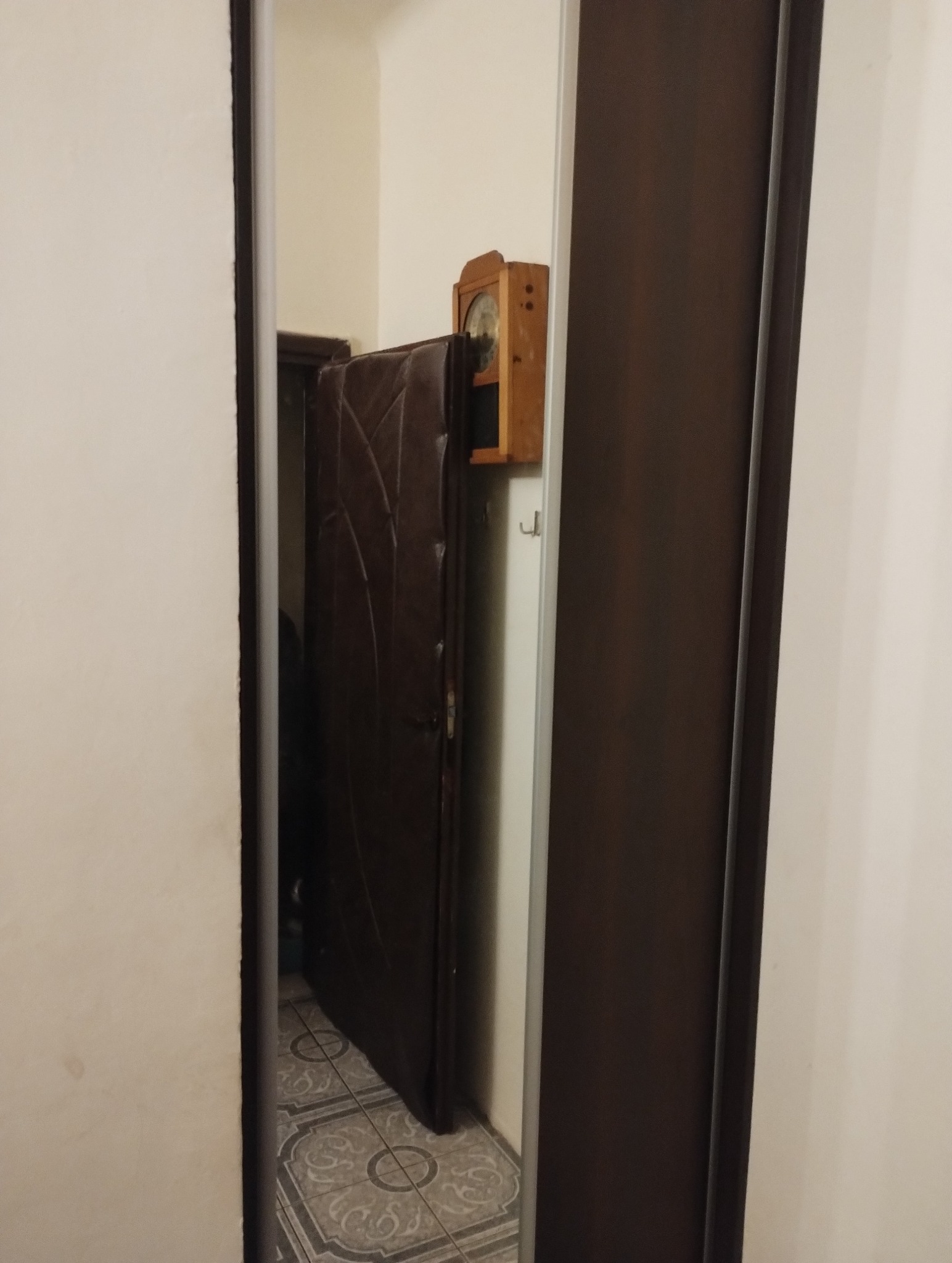 Ciemne drzwi wejściowe z dekoracyjnym wzorem widoczne z perspektywy otwartych drzwi wewnętrznych, na ścianie wisi drewniany zegar.