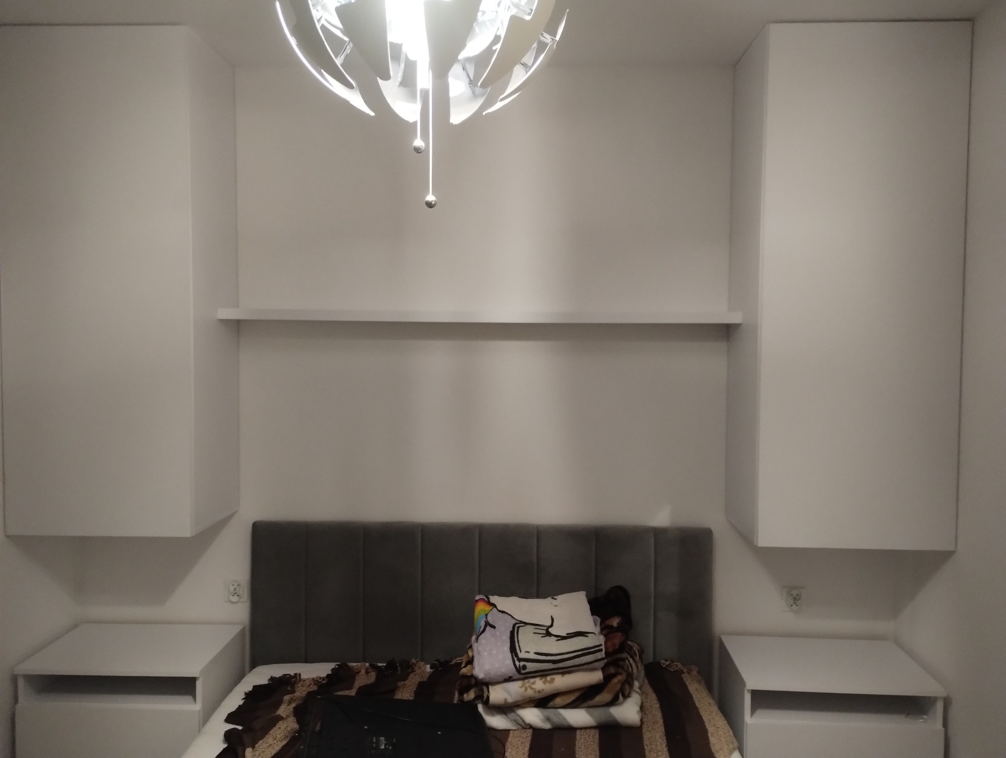 Nowoczesna sypialnia w Radomsku z białymi szafkami wiszącymi po bokach łóżka z szarym zagłówkiem, nad łóżkiem półka i designerska lampa.