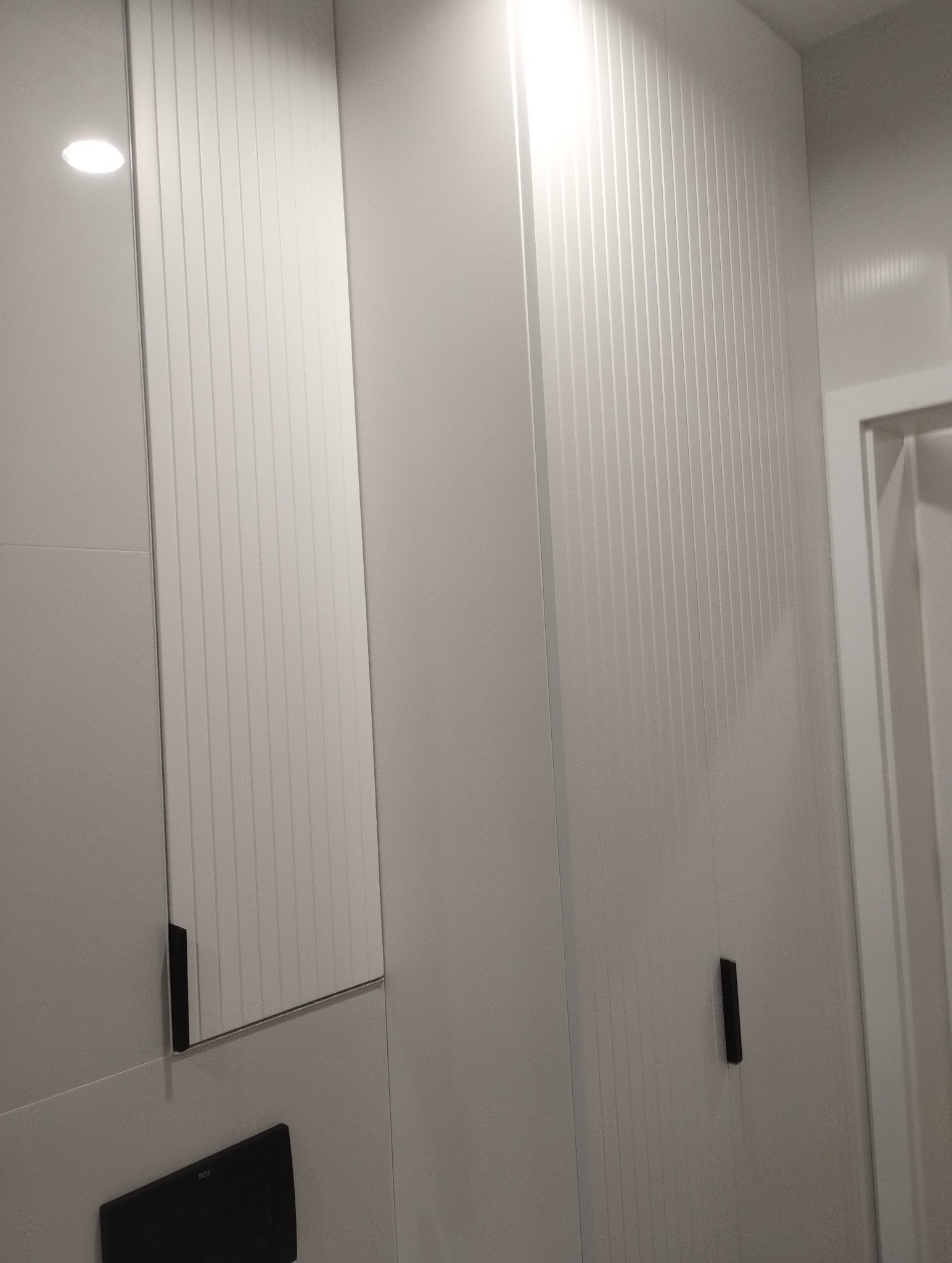 Biała szafa z ryflowanymi drzwiami i minimalistycznym uchwytem, obok lustro z czarnym elementem mocującym, fragment ściany i drzwi w tle.