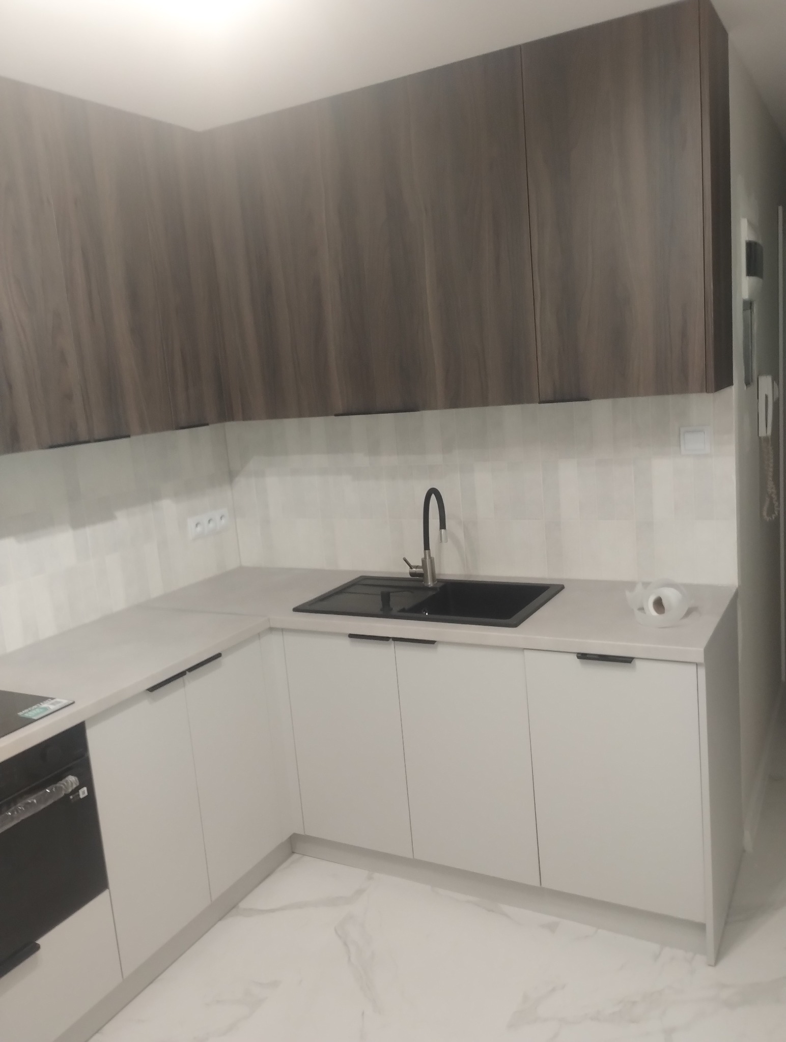 Nowoczesna kuchnia z białymi frontami szafek dolnych i górnymi szafkami w kolorze ciemnego drewna, czarnym zlewozmywakiem i armaturą, oraz białym blatem i białymi płytkami na ścianie.