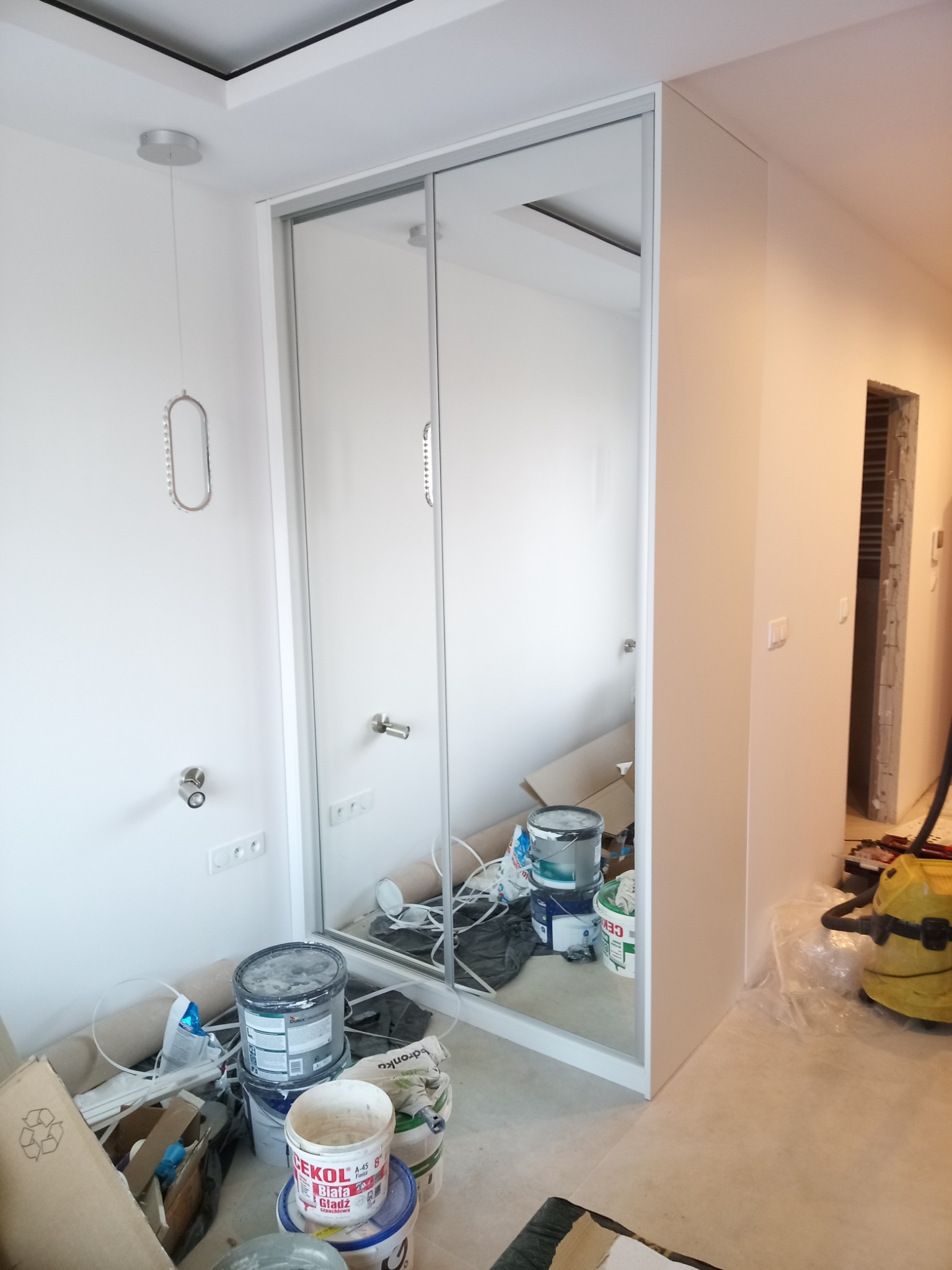 Biała szafa wnękowa z lustrzanymi drzwiami przesuwnymi w trakcie montażu, widoczne puszki z farbą i narzędzia malarskie, wnętrze w trakcie wykańczania.