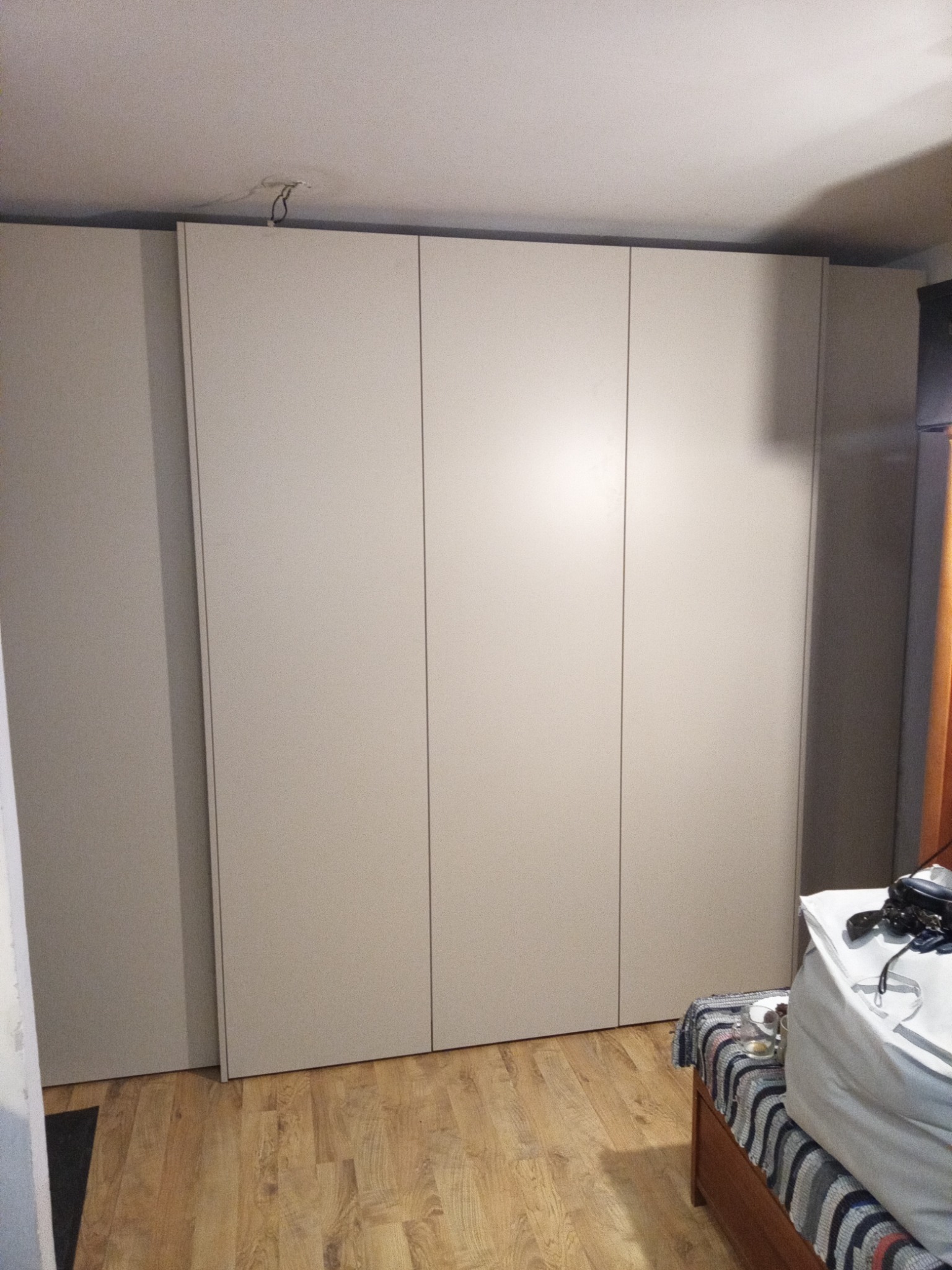 Pełna ściana z zabudowaną, trzydrzwiową szafą w odcieniach beżu, z widocznymi kablami wystającymi z sufitu i fragmentem łóżka z pościelą w prawym dolnym rogu.